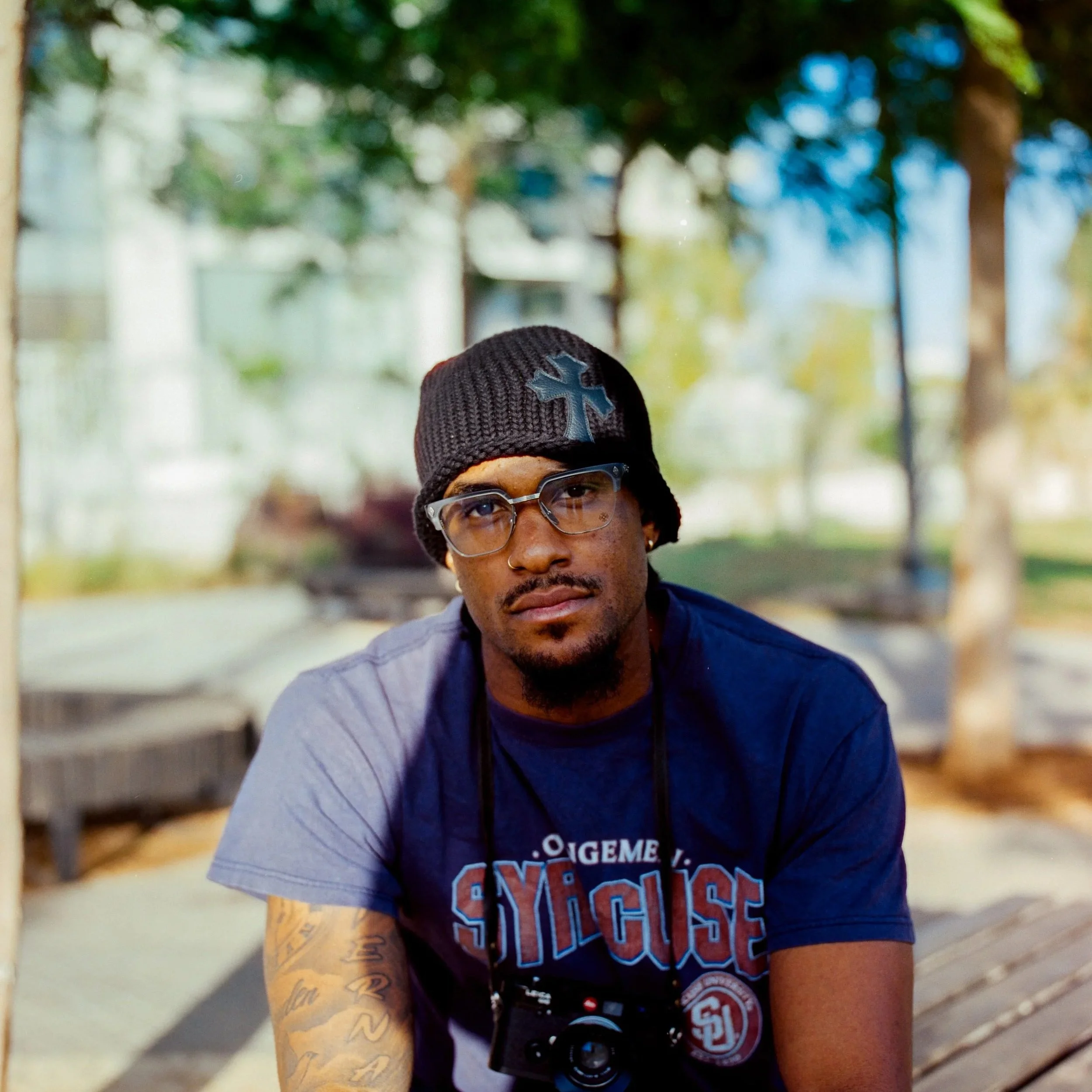 A man wearing a black beanie, glasses, and a navy blue T-shirt with a Syracuse logo, sitting outdoors on a wooden bench with trees and a blurred background.