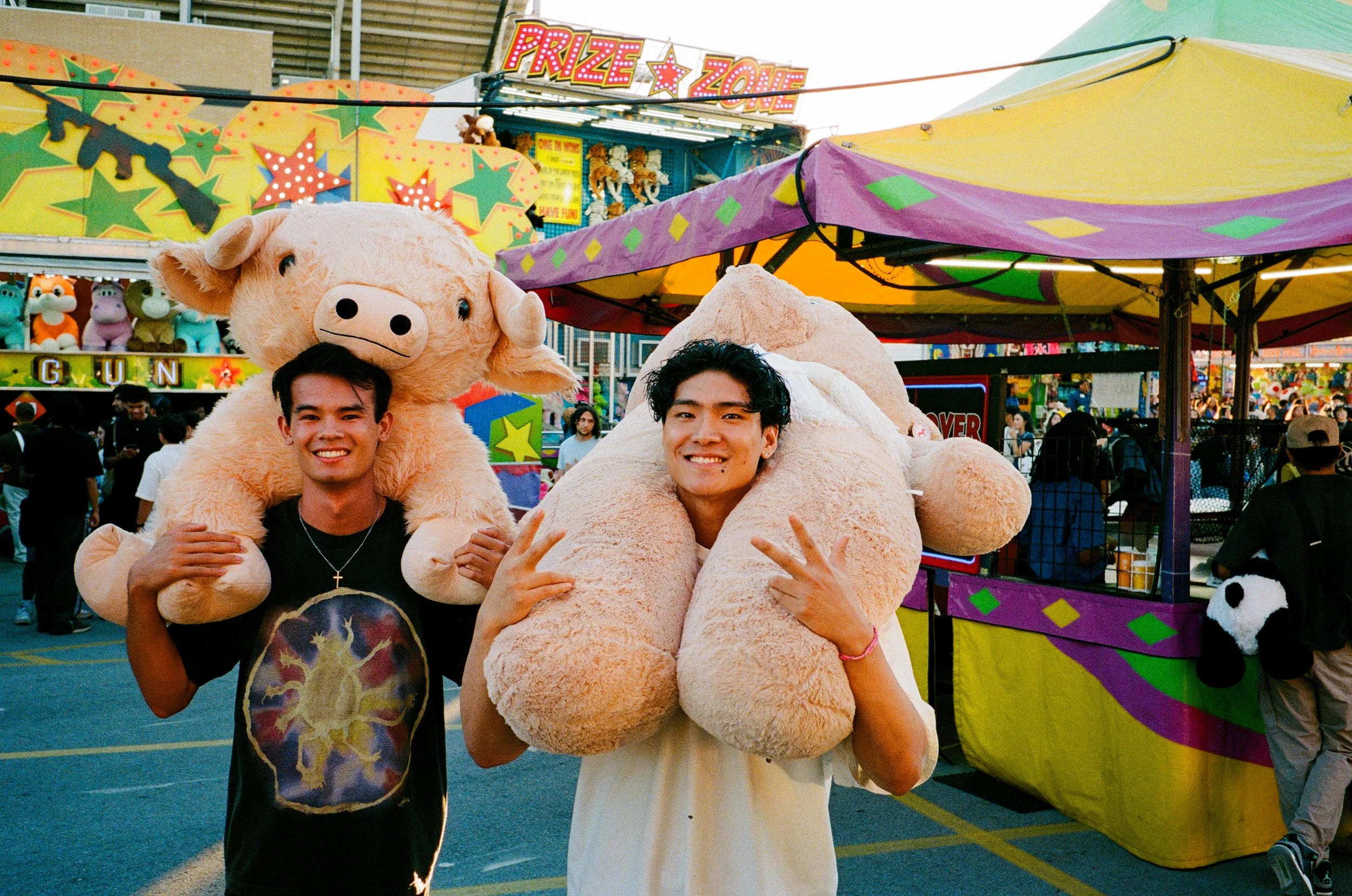 Two young men holding large plush pig and sheep toys on a colorful amusement park ride with game booths and carnival tents in the background.