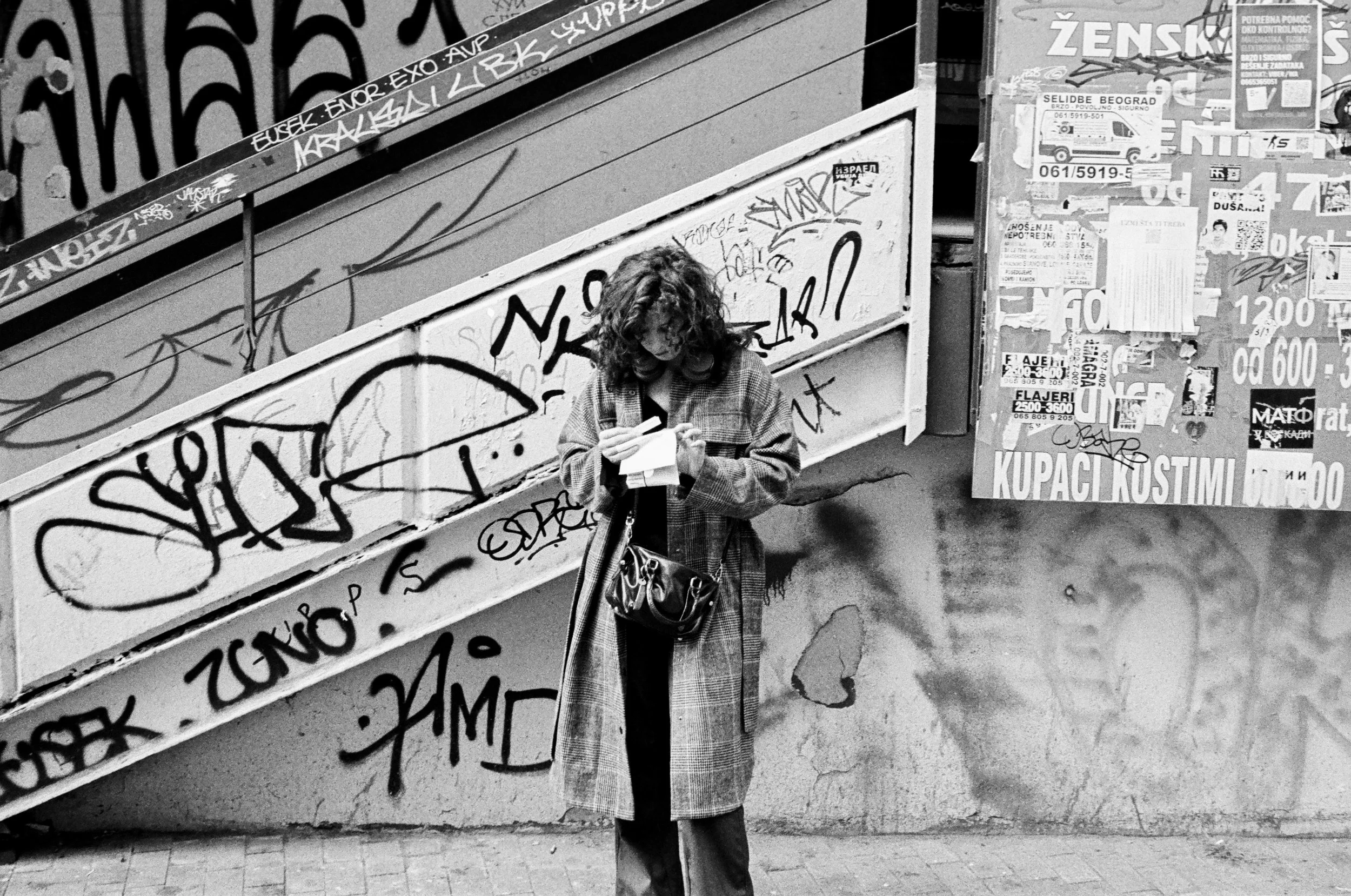 A woman with curly hair wearing a long plaid coat, looking at her phone while holding a small shopping bag, standing on a sidewalk near a graffiti-covered staircase and a bulletin board with posters.