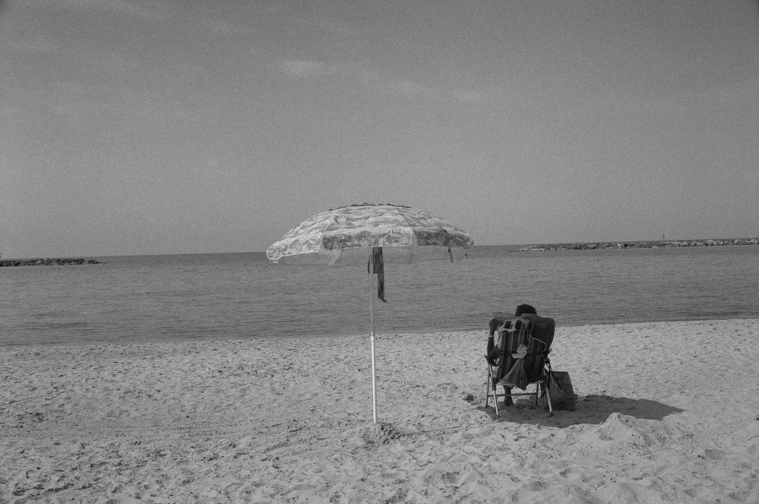 A person sitting on a beach chair under an umbrella, facing the ocean with rocks in the distance on a sandy beach.