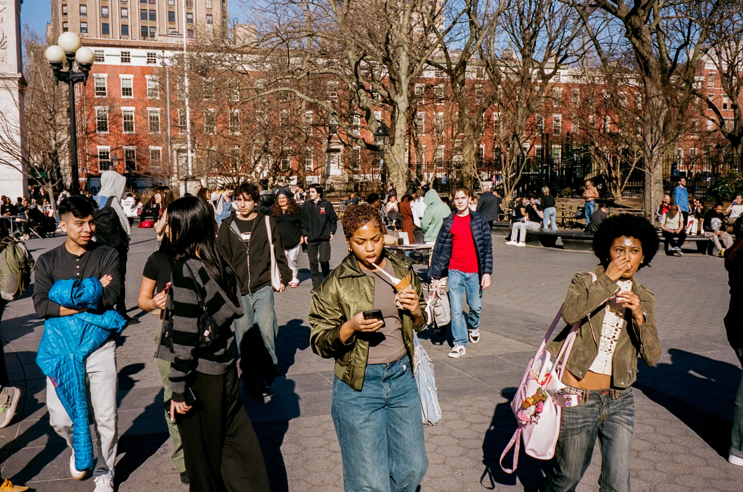 People walking and sitting in a park on a sunny day, with leafless trees and red brick buildings in the background.