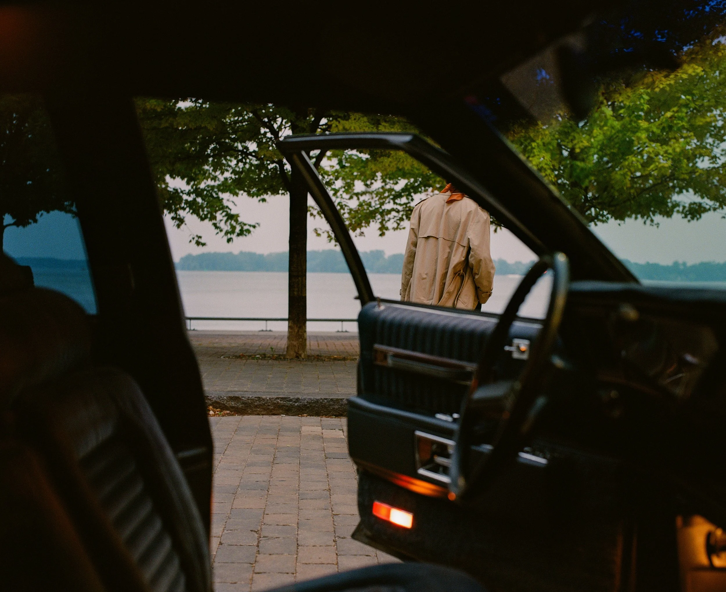 View from inside a vehicle looking out at a person standing near a lake, partially obscured, with trees and pathway in the background.