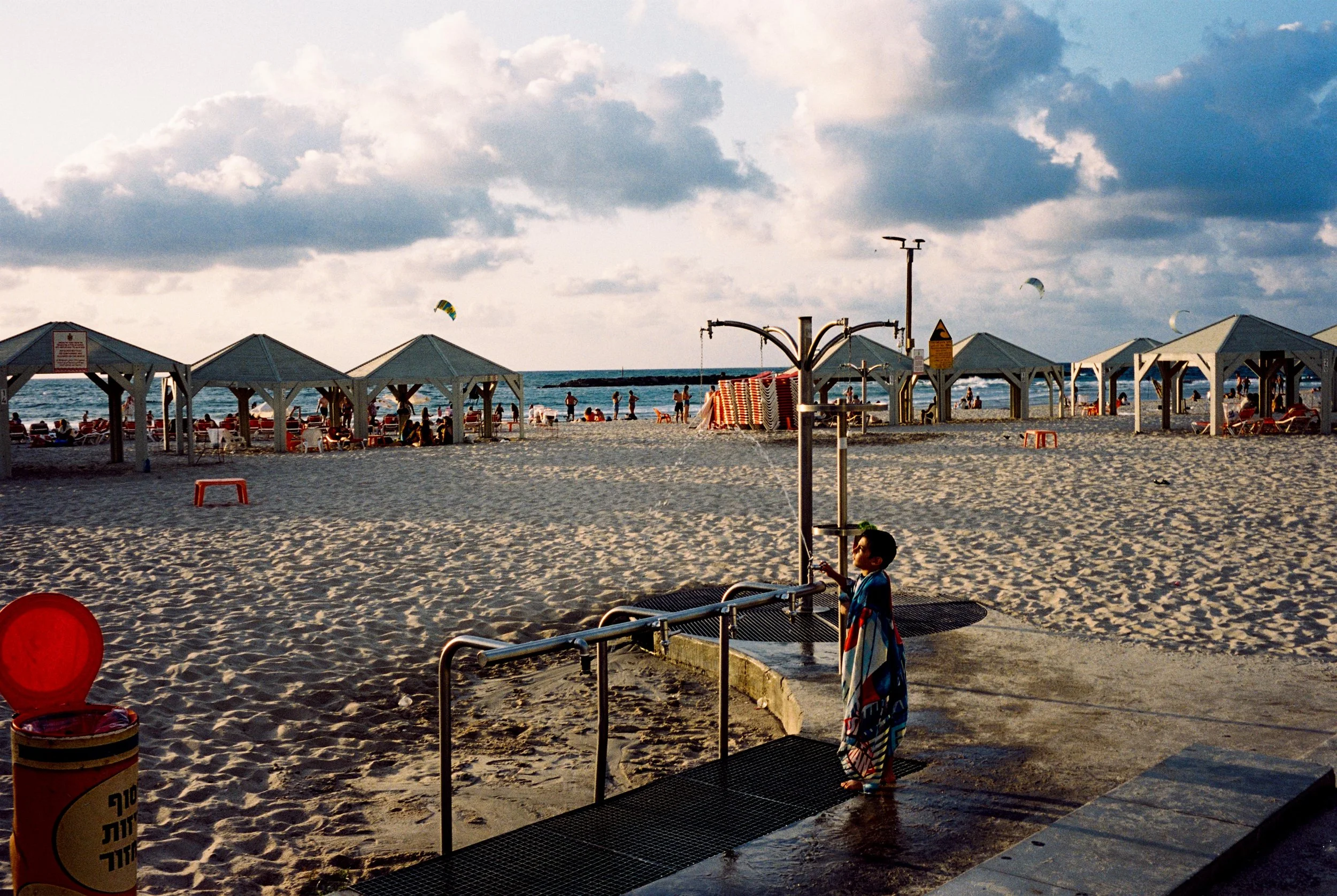 A child standing at an outdoor shower on a sandy beach, with beach huts and people relaxing by the water in the background. The sky is partly cloudy.