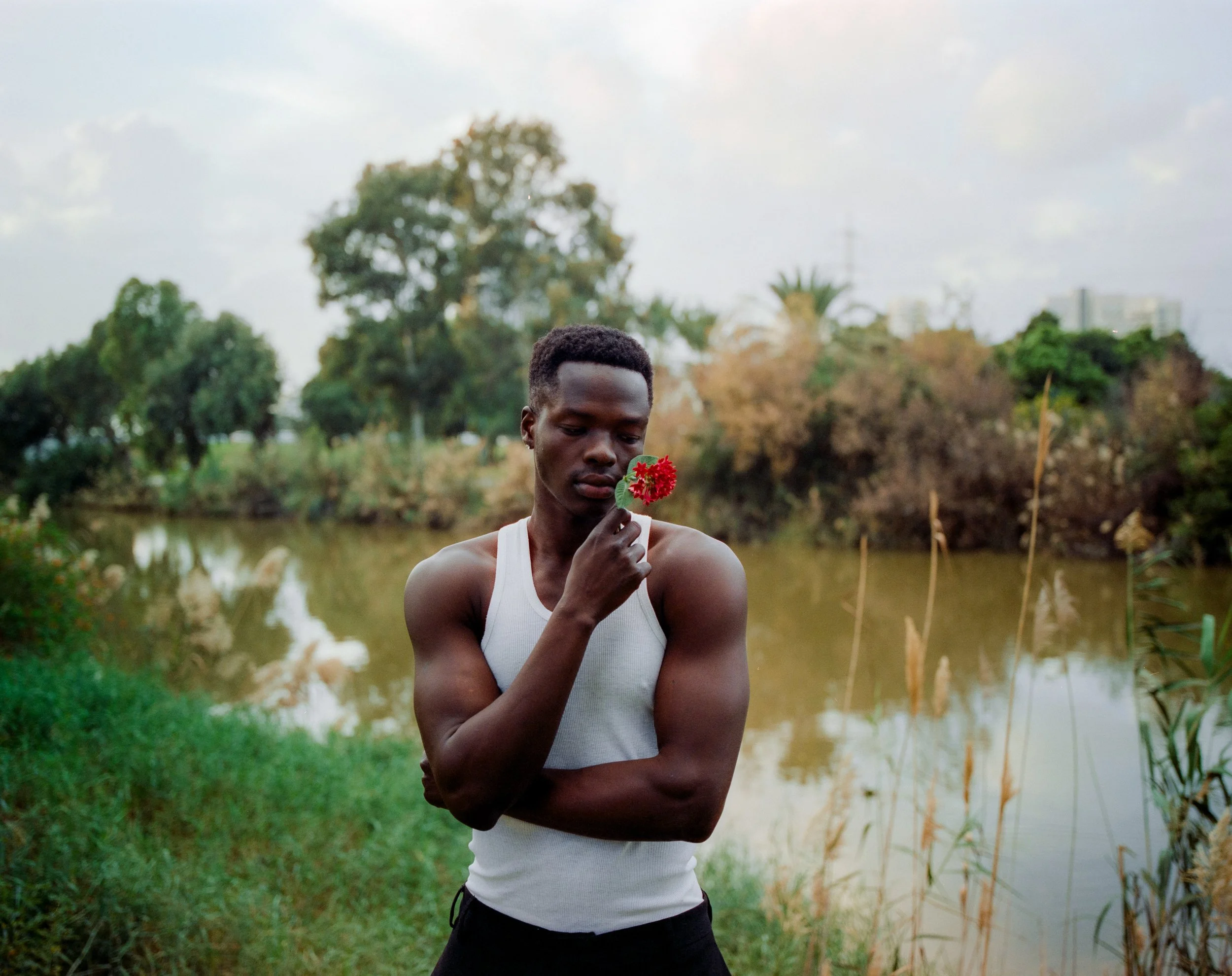 A young man with dark skin, wearing a white tank top, stands by a body of water holding a red flower near his face, with trees and a cloudy sky in the background.