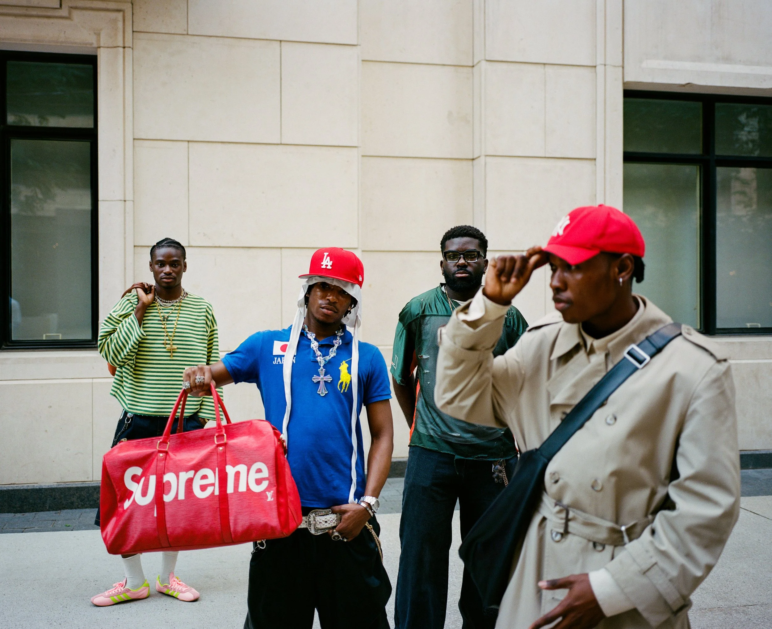 Four young men standing on a city sidewalk in front of a beige building. One is holding a large red Supreme duffel bag. They are dressed in colorful streetwear, with two wearing baseball caps and one wearing sunglasses.