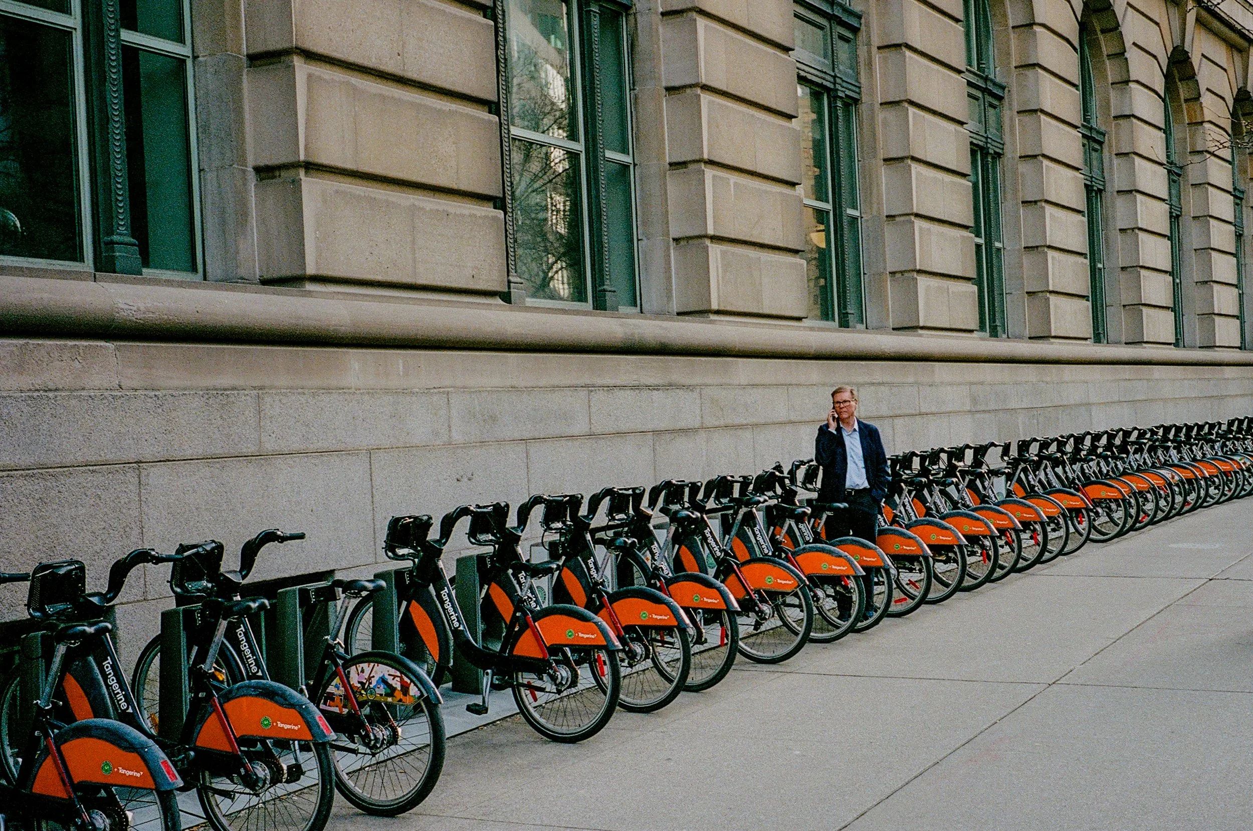 A man in a business suit is standing on a sidewalk next to a row of orange and black community bikes parked at a docking station, with a large stone building with tall windows in the background.