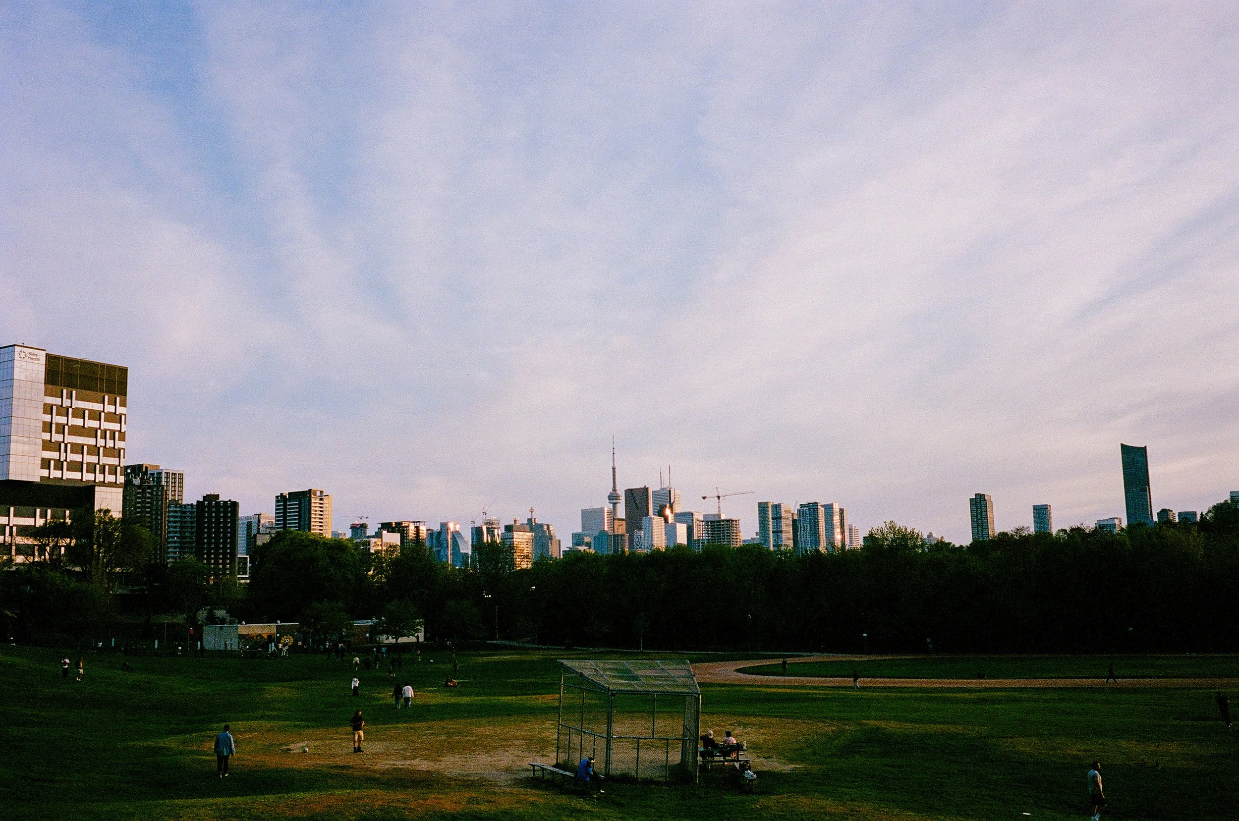 A city skyline with tall buildings and a tower, seen from a park with green grass and people walking or sitting underneath a small shelter.