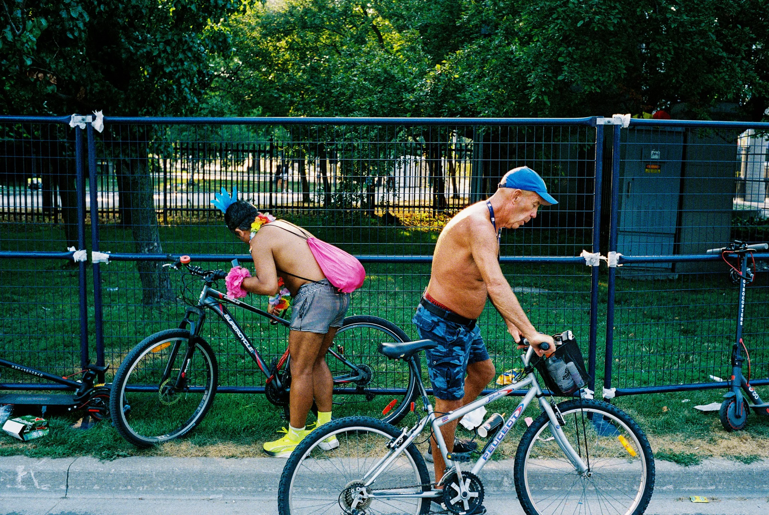 Two shirtless men with bicycles stand next to a blue metal fence in a park. One man is wearing a blue cap, shorts, and is riding a bike. The other man is wearing sunglasses, a yellow glove, shorts with a pink cloth, and has a colorful feathered mask 