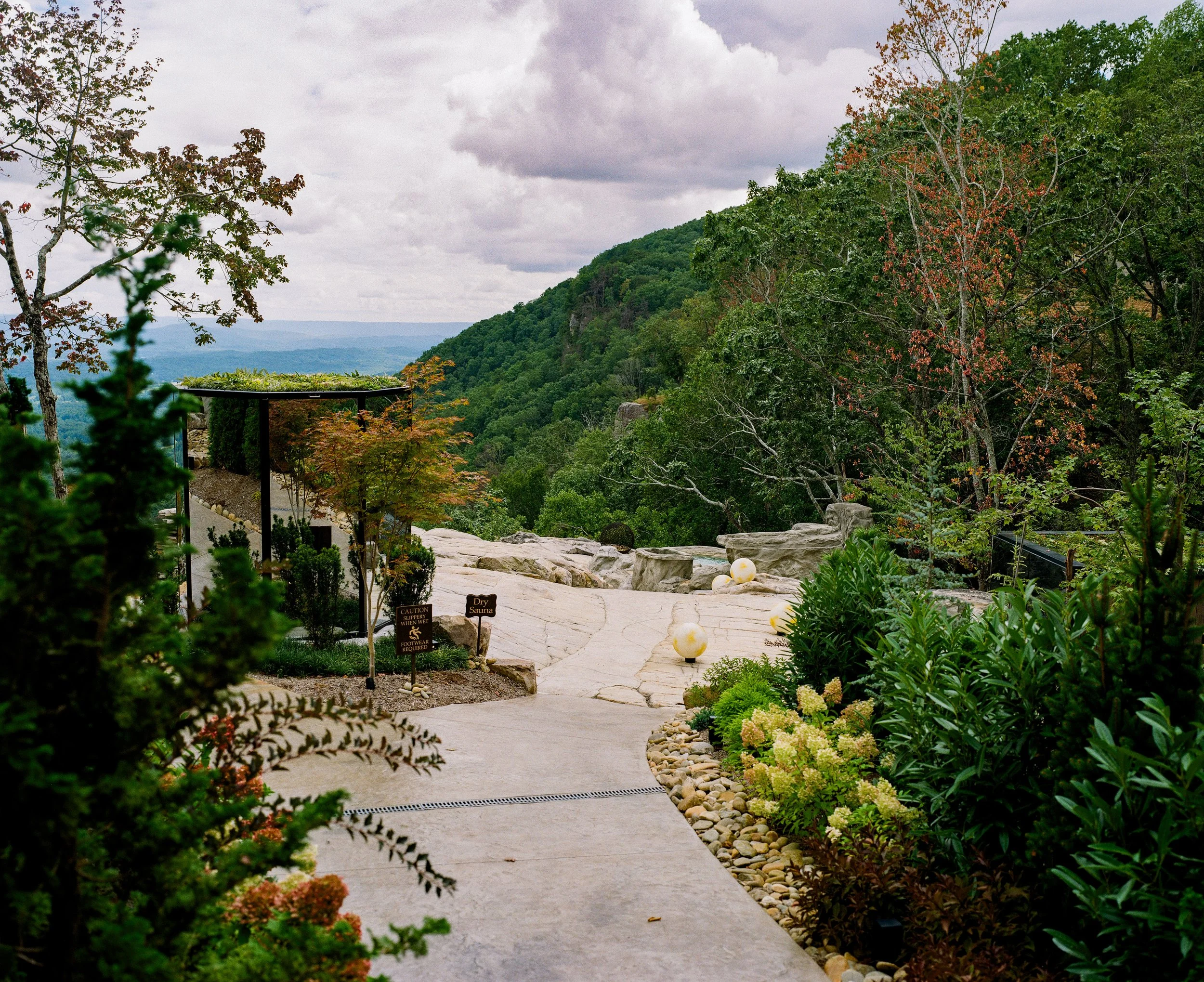 Landscape with lush green trees, a paved pathway, large rocks, and white spherical lights, overlooking a valley with distant mountains and cloudy sky.
