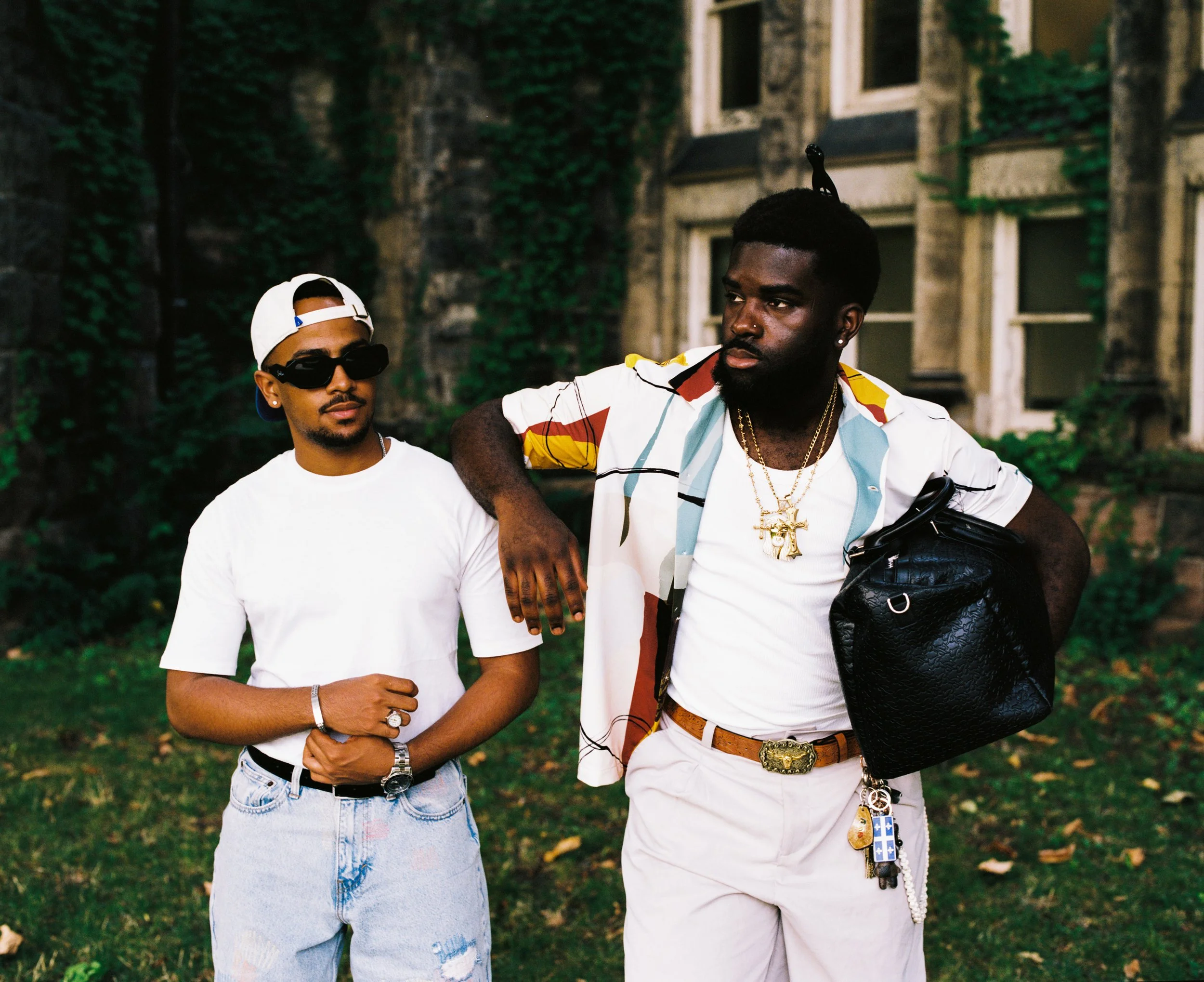 Two young men standing outdoors in front of a brick building covered in green vines. One is wearing a white t-shirt, distressed jeans, sunglasses, and a backwards white cap. The other is wearing a colorful short-sleeve shirt over a white t-shirt, bei