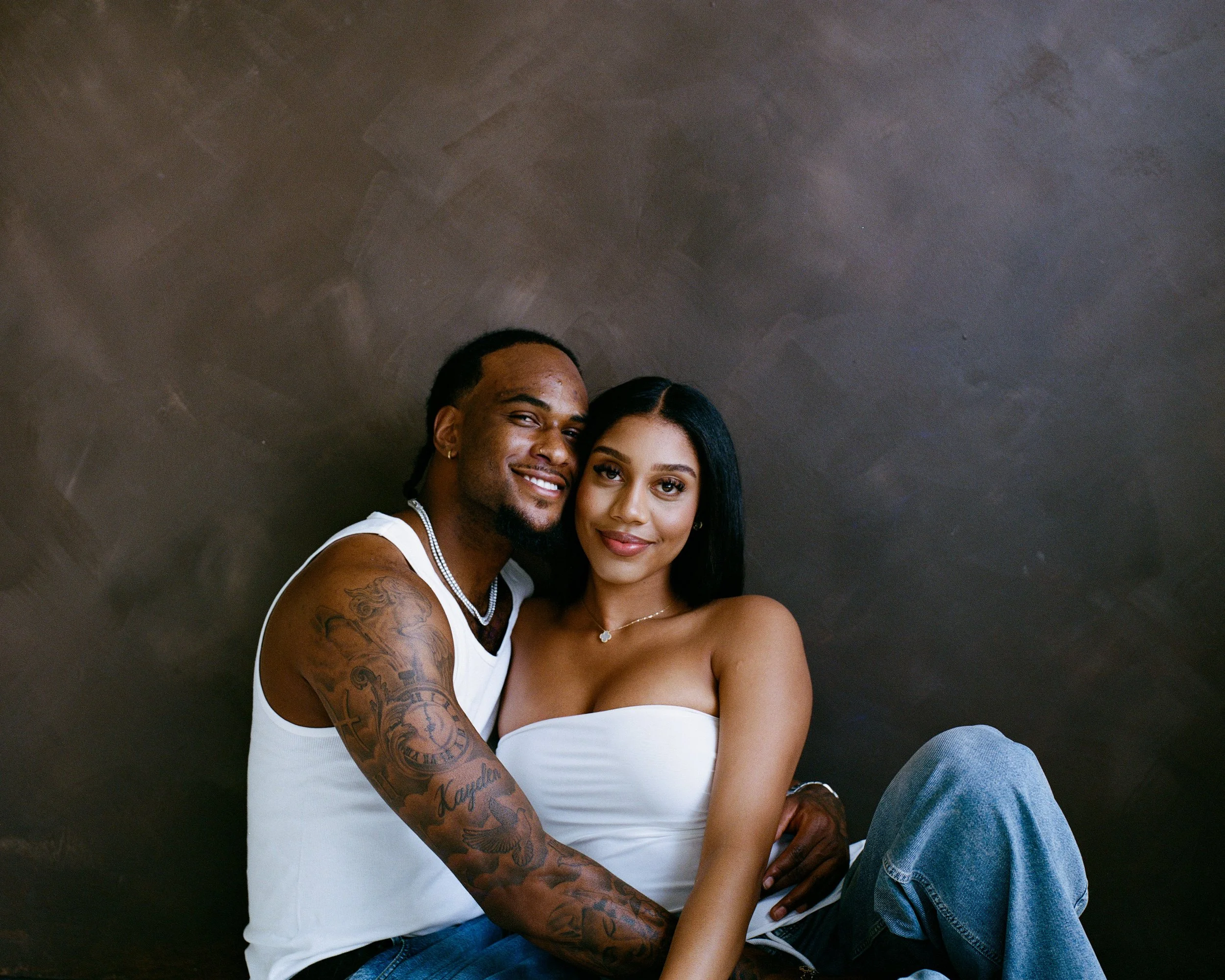 A happy couple sitting close together, smiling at the camera, with a dark textured wall background.