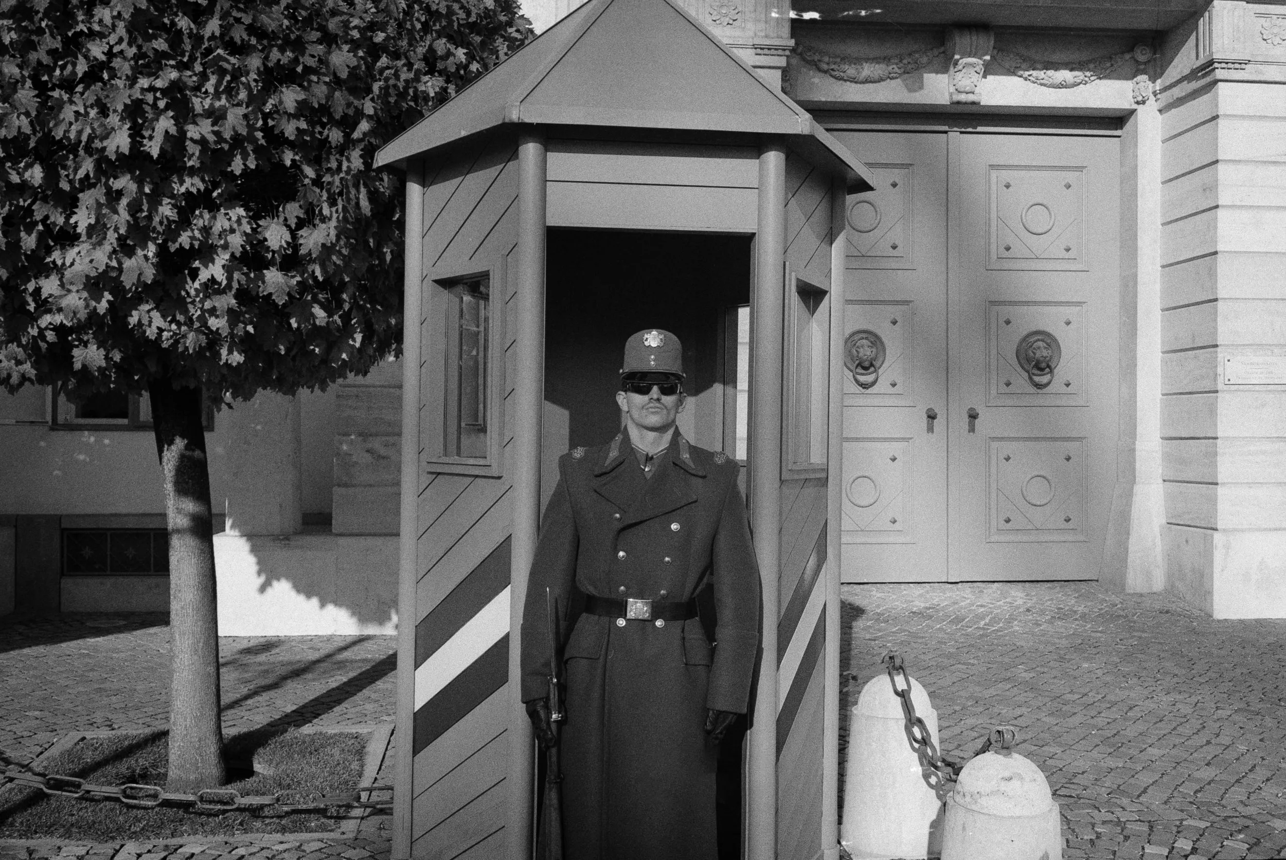A uniformed soldier standing inside a small guard booth, wearing sunglasses and a cap with a badge, in front of a historic building with ornate architecture and large decorative doors.