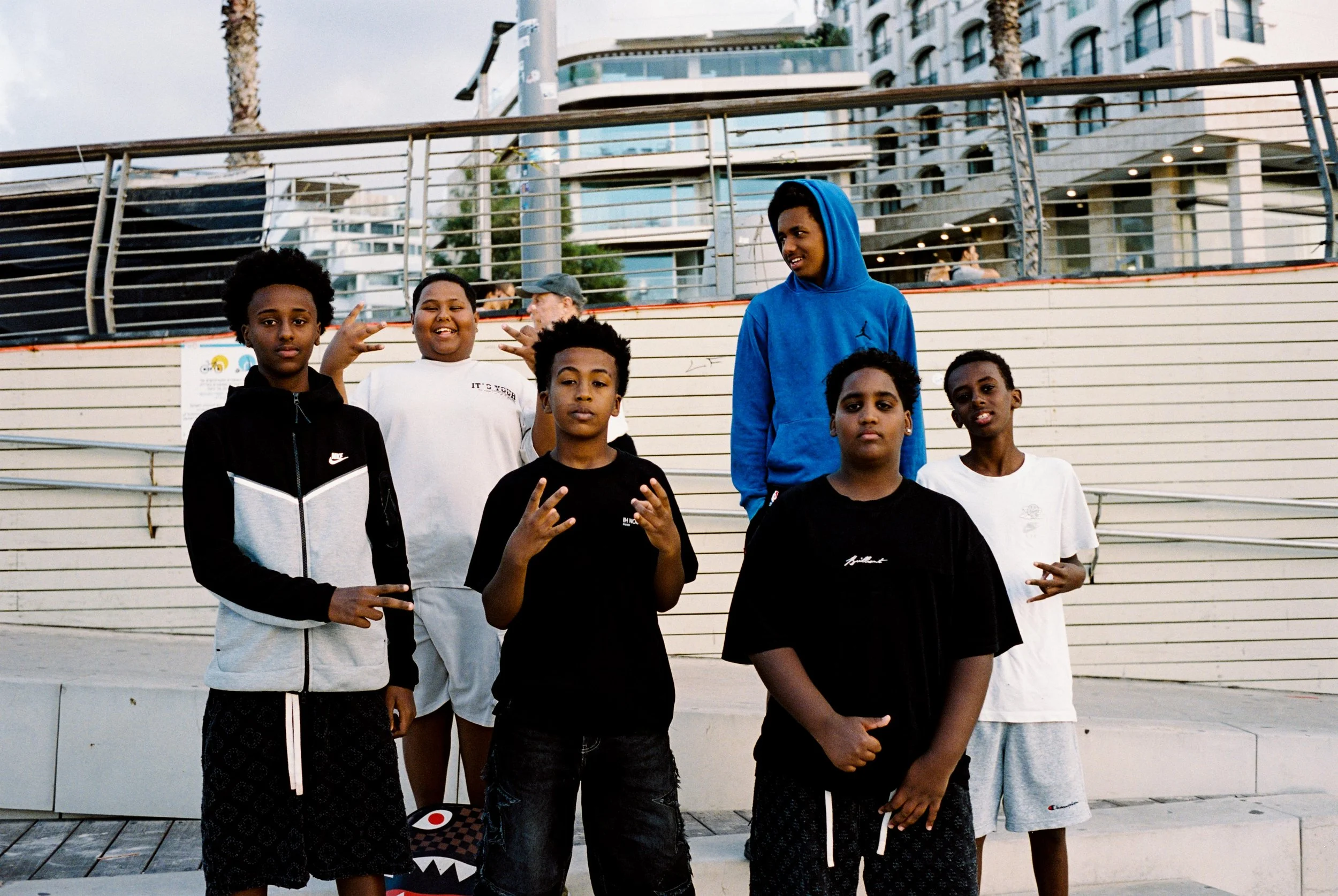 Group of six boys standing outdoors on steps near a railing, with modern buildings and palm trees in the background.