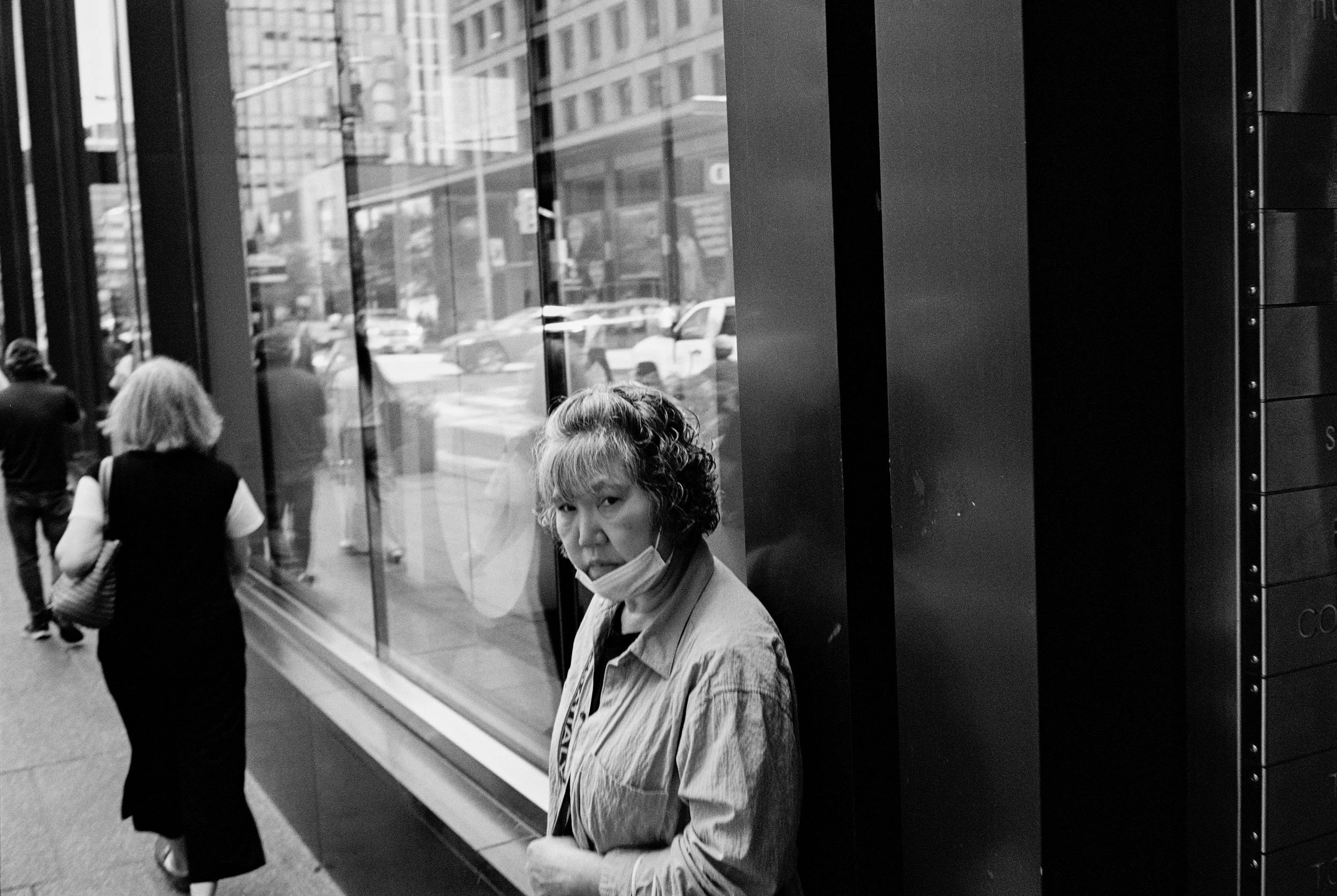A woman with curly hair and a face mask hanging below her chin, standing beside a glass building in an urban setting with cars and pedestrians in the background.