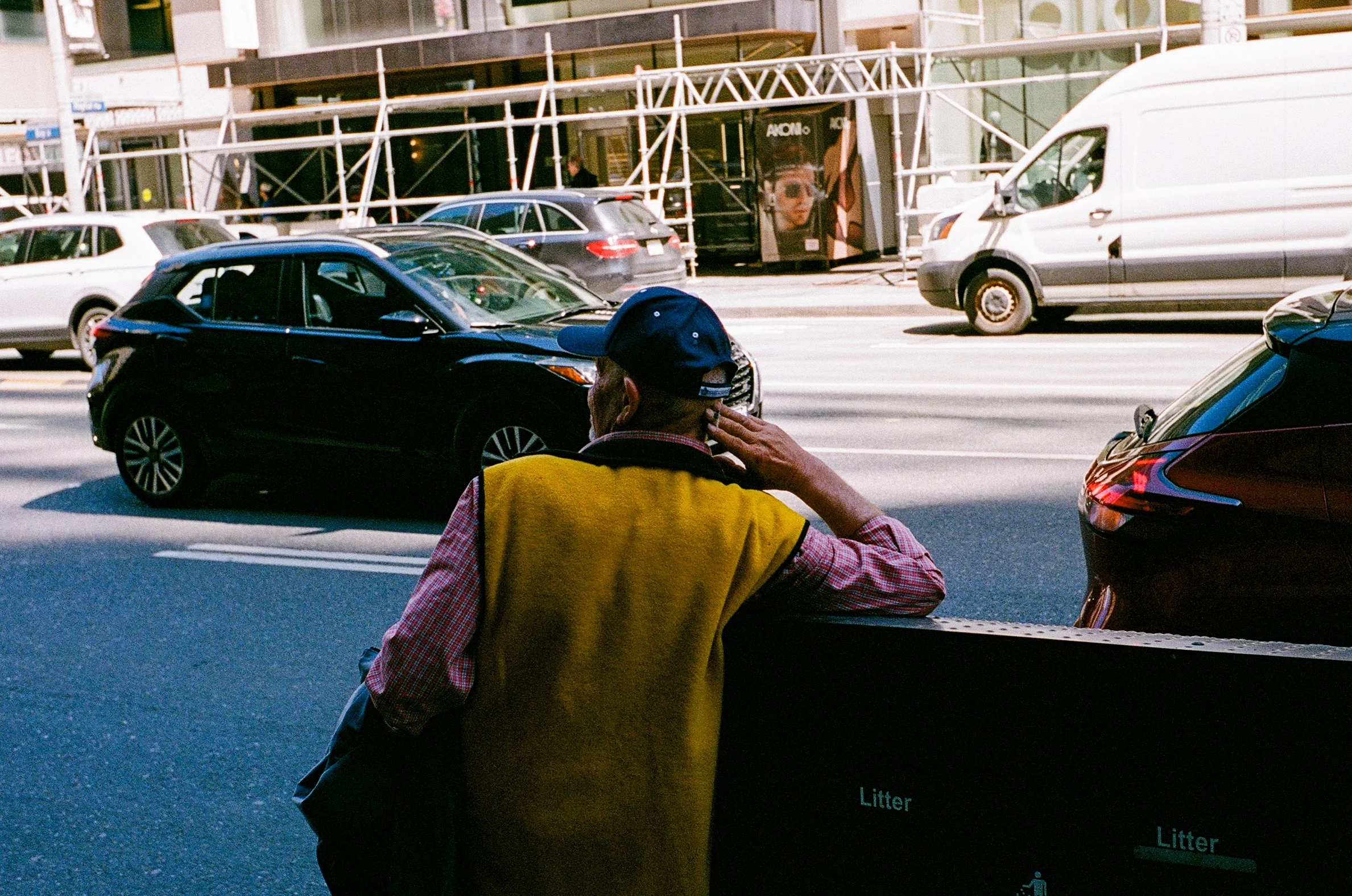 An elderly man wearing a yellow vest and a blue cap stands on a city sidewalk, leaning on a black litter bin, looking across the street at parked and moving cars, with a construction site and buildings in the background.