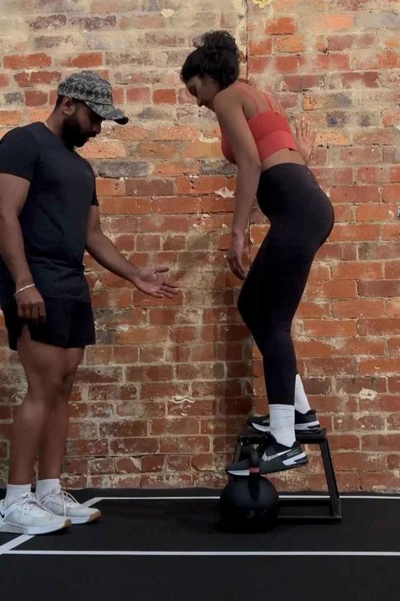 A woman balancing on a workout bench with a kettlebell underneath her feet, assisted by a trainer standing nearby.
