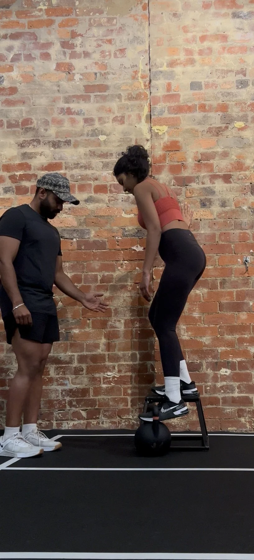 A woman balances on a black workout step with her feet on a medicine ball, while a trainer stands nearby, giving her instructions, in a gym with a brick wall background.