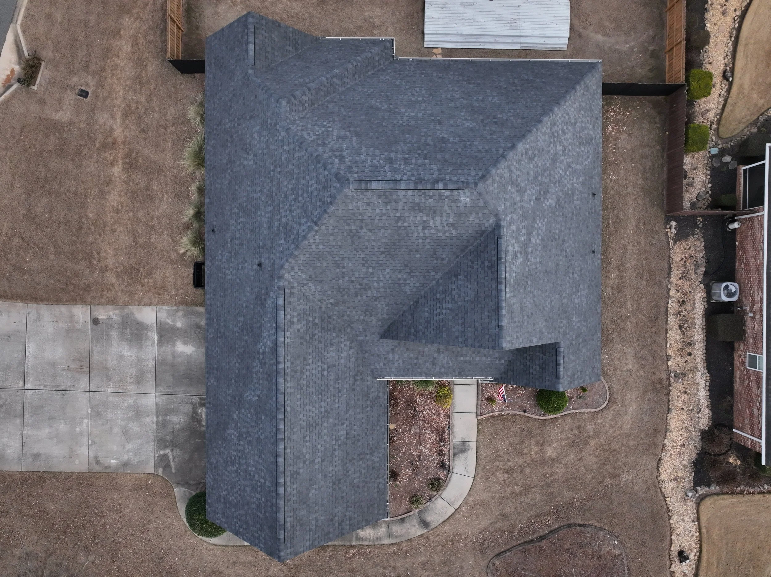 Top-down aerial view of a house with a gray shingled roof, surrounded by a concrete driveway, patches of dry grass, and a landscaped area with small bushes and a sidewalk.