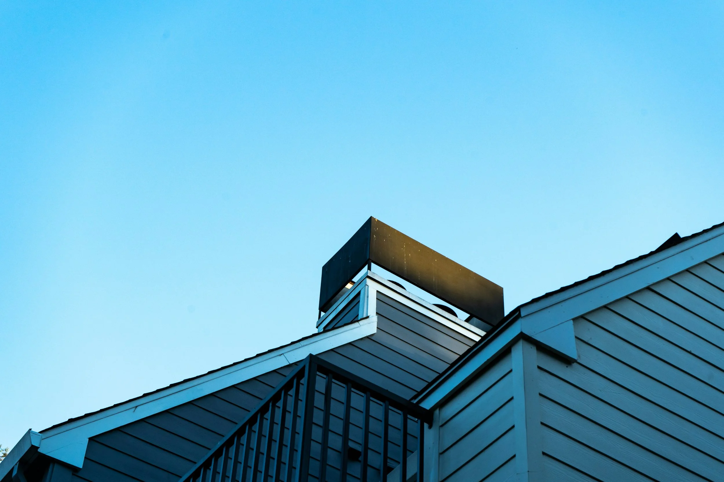 The top of a building with dark siding and white trim, featuring a chimney or vent on the roof, against a clear blue sky.