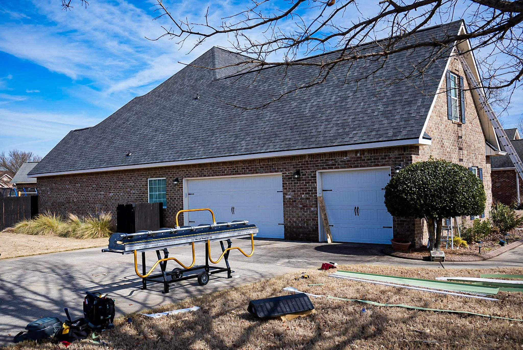 A residential garage with two white doors and a brick exterior. Construction tools and equipment are on the driveway and yard, indicating ongoing work. A neatly trimmed bush is on the right side of the garage, with a ladder leaning against the house.