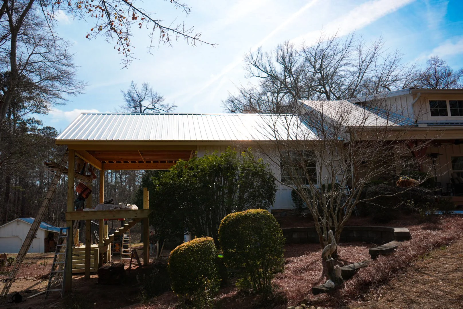 A house with a metal roof under construction, surrounded by trees, bushes, and a landscaped yard on a sunny day.
