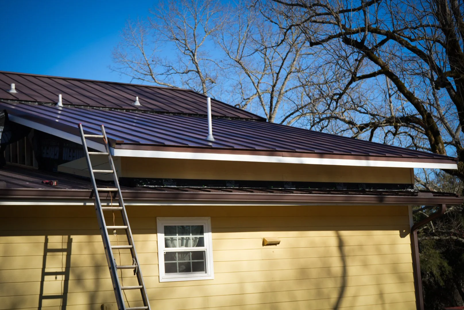 A house with light yellow siding undergoing roof construction, with a ladder leaning against the house. The roof has new brown metal panels and some construction details visible. Bare trees are in the background under a clear blue sky.