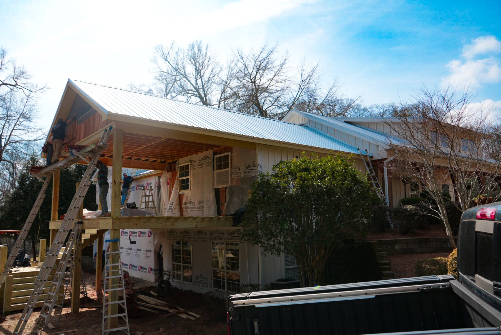 Construction workers building a house with a metal roof, using ladders and scaffolding, surrounded by trees on a sunny day.