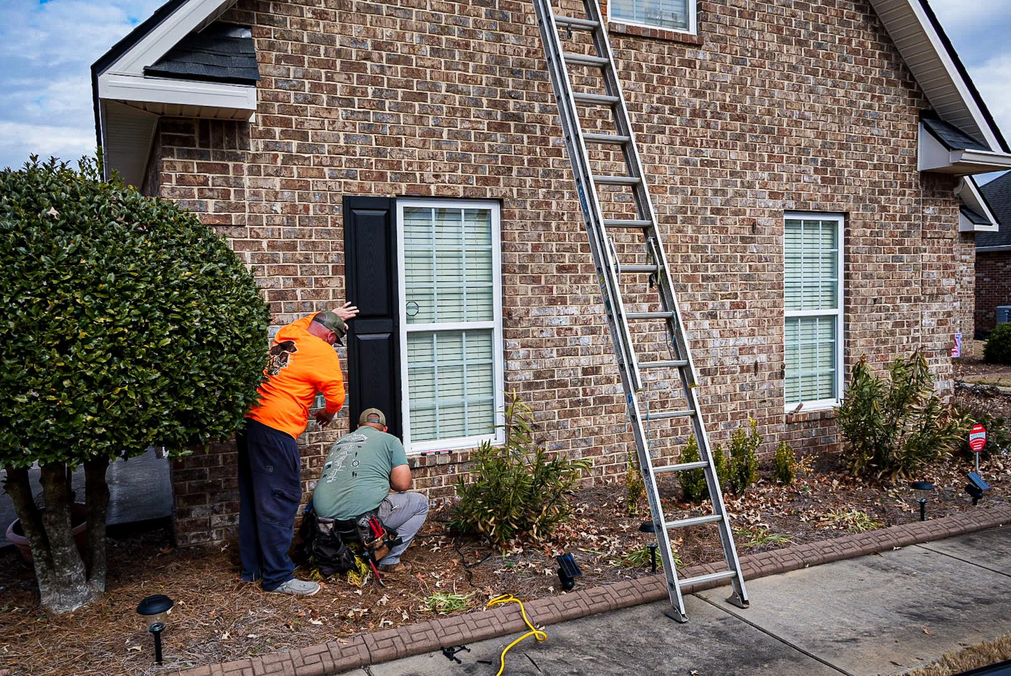 Two workers installing or repairing a black window shutter on a brick house. A silver ladder leans against the house, and there are small landscape lights, bushes, and a concrete walkway in the front yard.