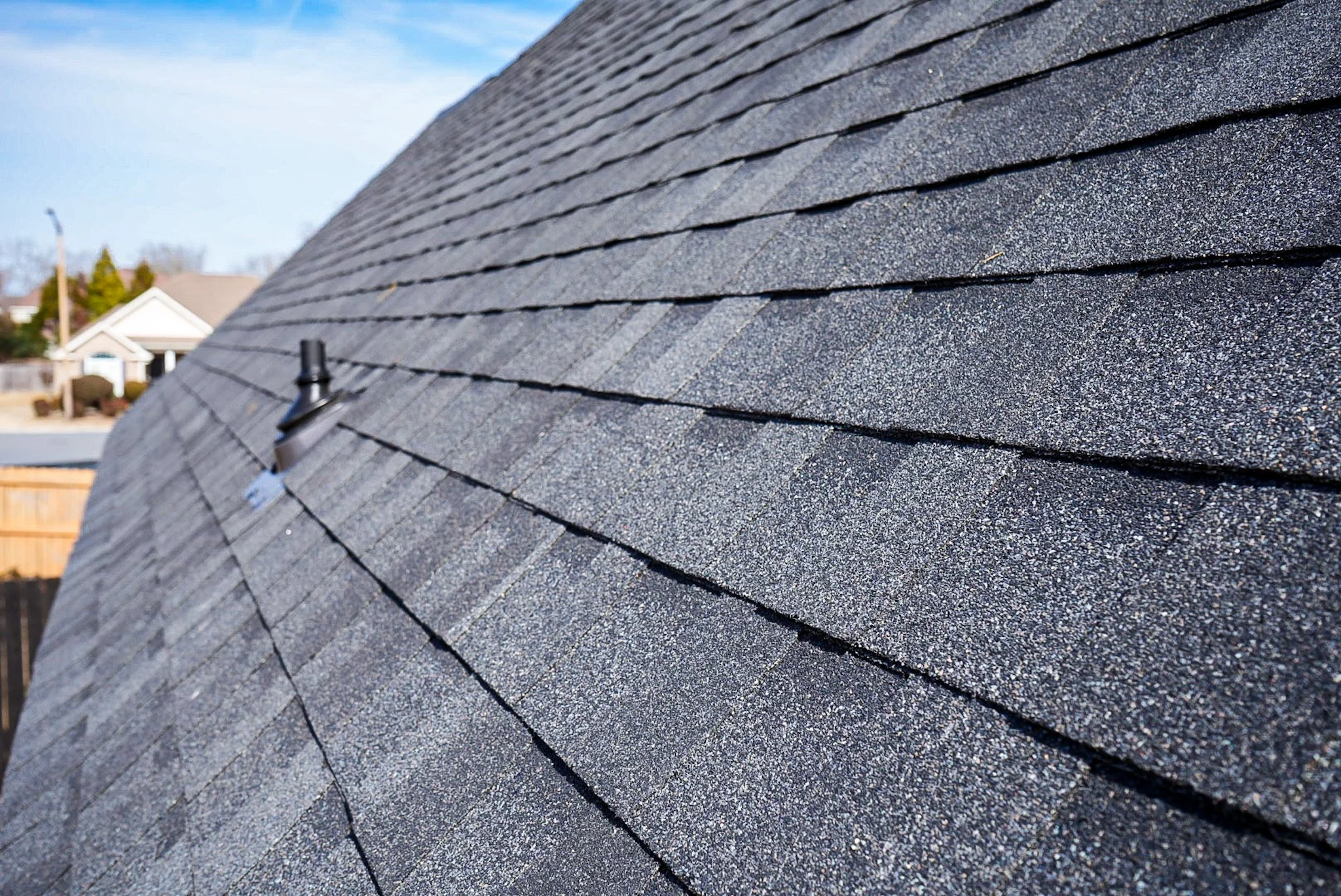Close-up view of a black asphalt shingle roof on a house, showing textured, layered shingles extending into the distance, with a chimney pipe visible.