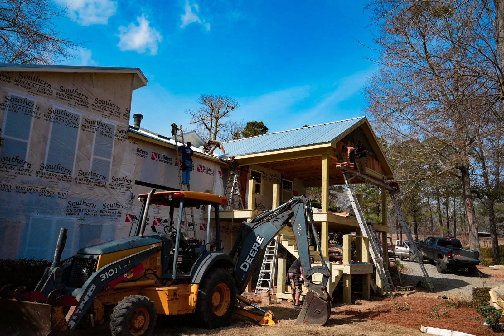 Workers are constructing a house with a blue metal roof. Some are on the roof, others are on ladders. A construction vehicle is parked in front of the house. The house has wooden framing, and there are trees and parked cars in the background.