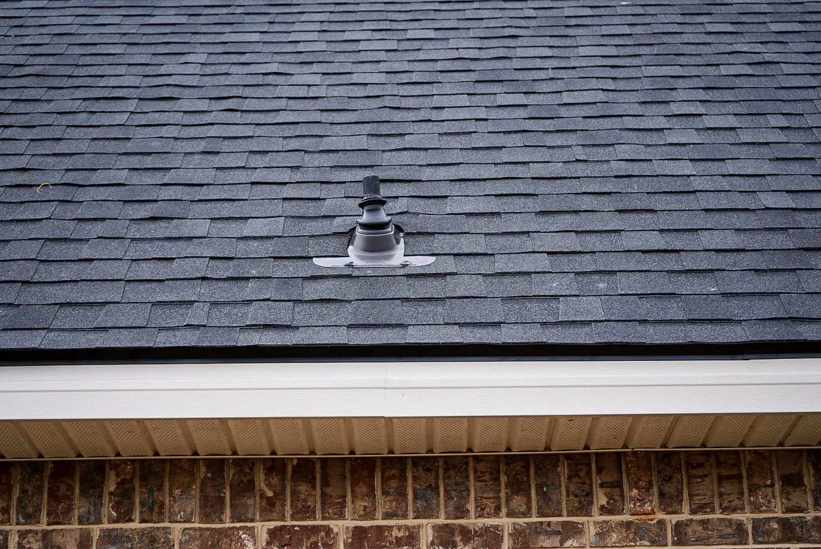 Close-up of a house roof with dark gray shingles, a roof vent pipe, white trim, and brick wall at the bottom.