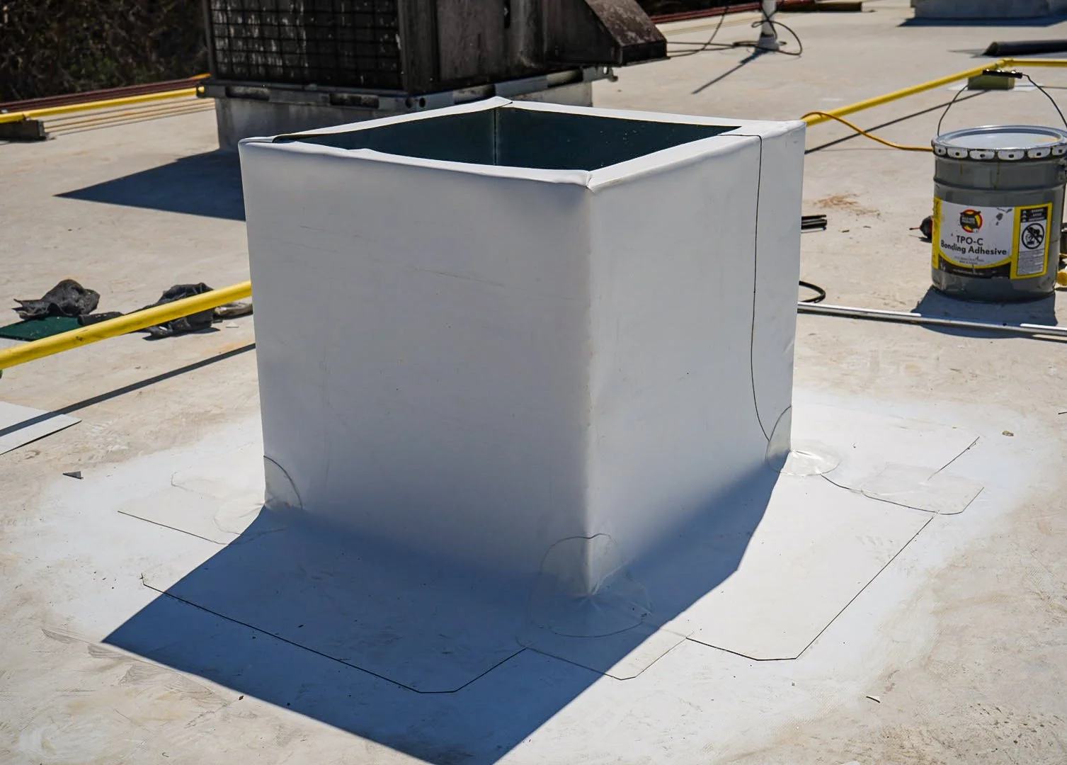 A square metal vent pipe on a building rooftop, surrounded by construction materials and tools, including a bucket of bonding adhesive, black gloves, yellow electrical conduits, and electrical wires.