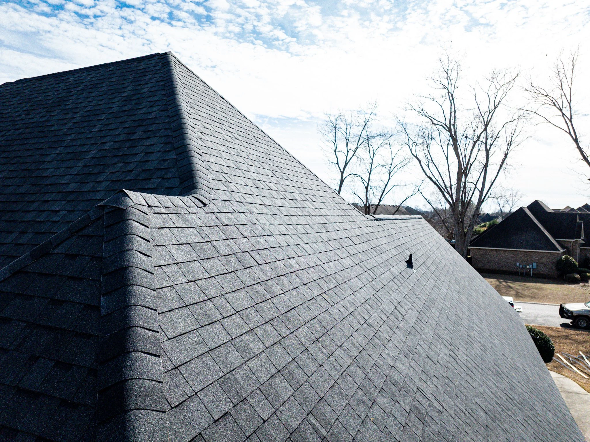 Close-up of a house roof covered with gray asphalt shingles; trees and houses in the background, partly cloudy sky.