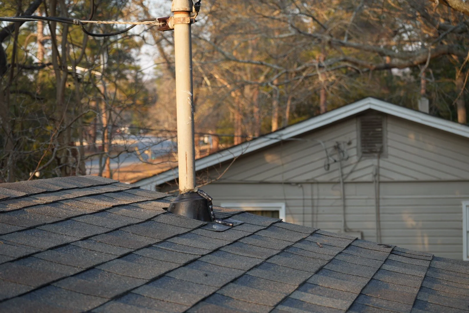 View of a house roof with asphalt shingles, a chimney pipe protruding from the roof, and a neighboring house in the background with trees and a clear sky.