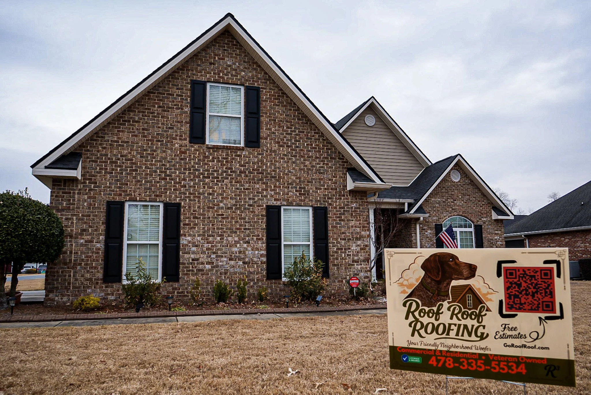 A two-story brick house with black shutters on the windows and a landscaped front yard. In the yard, there is a sign advertising roofing services, featuring a dog illustration and a QR code.