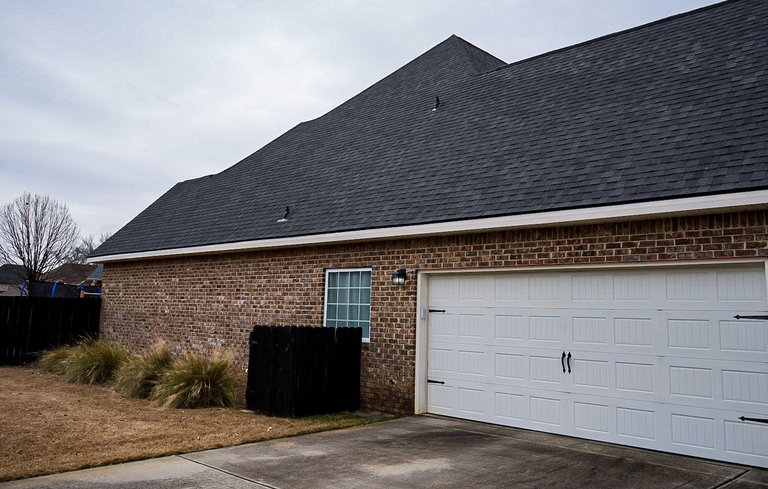 The side of a brick house with a white garage door and a window, surrounded by a small yard with grass and plants, and a black wooden fence.