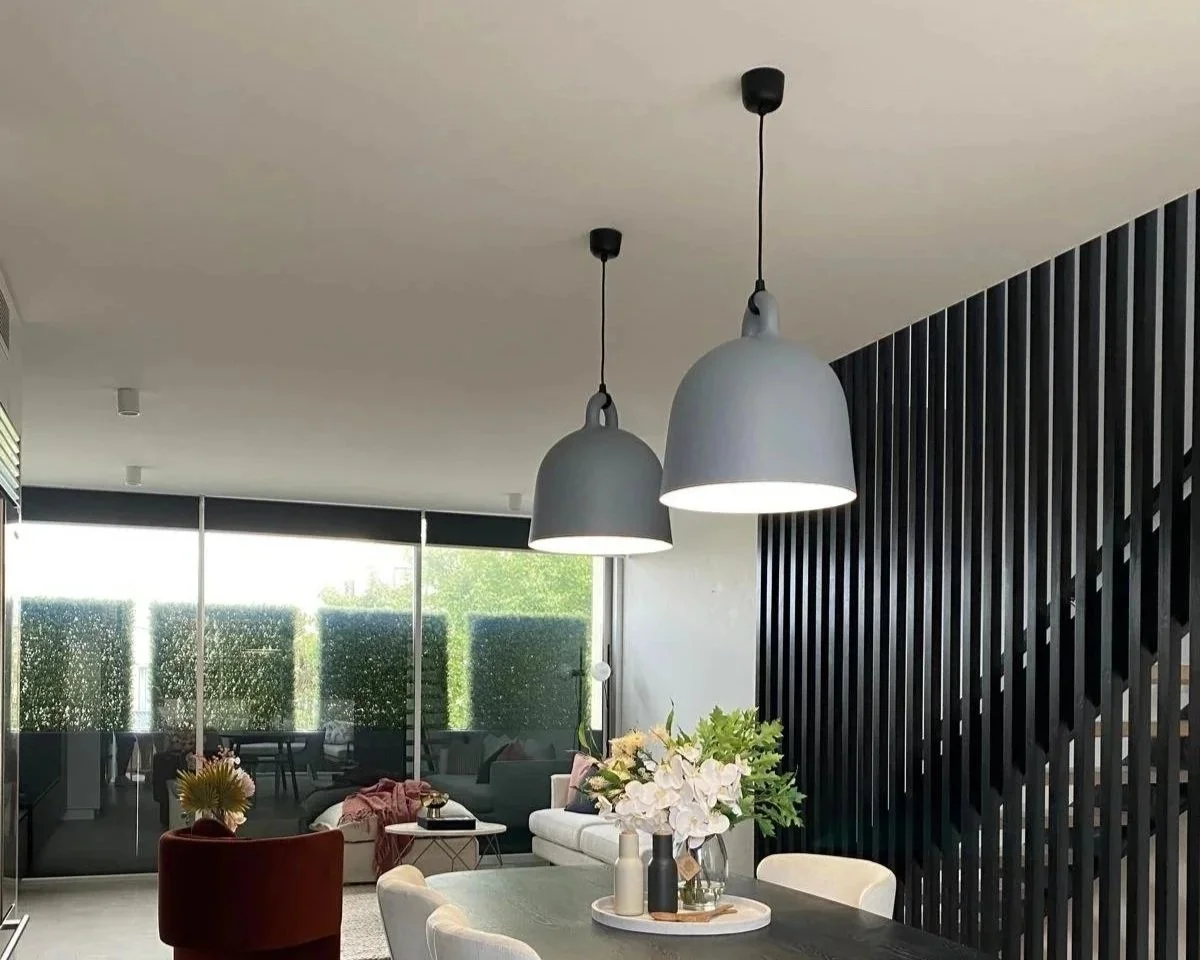Modern dining room with a black wooden partition wall, hanging pendant lights, white chairs, a gray table with a vase of white flowers, a white sofa, and large glass windows with outside greenery.