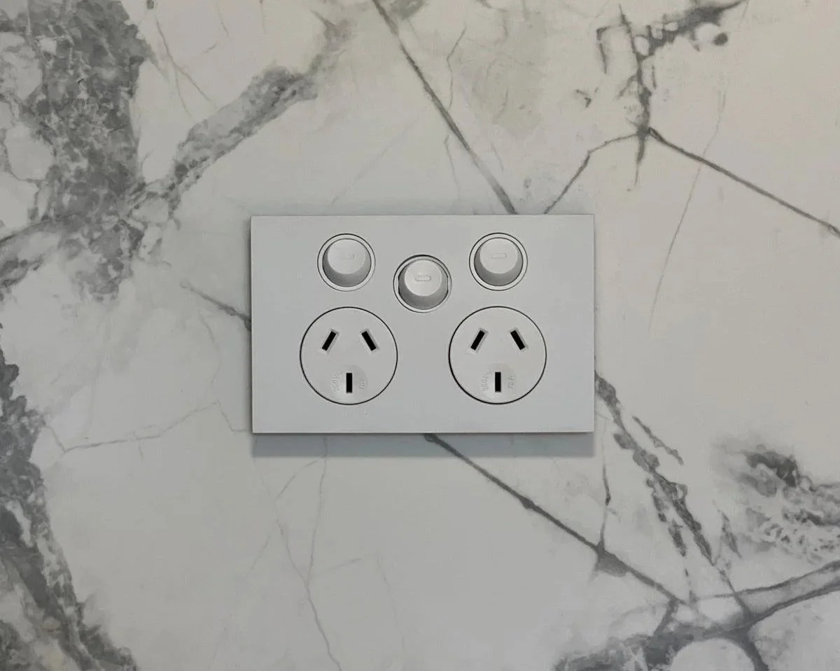 A white electrical outlet panel with two American-style power sockets and three small round switches, mounted on a marble wall.