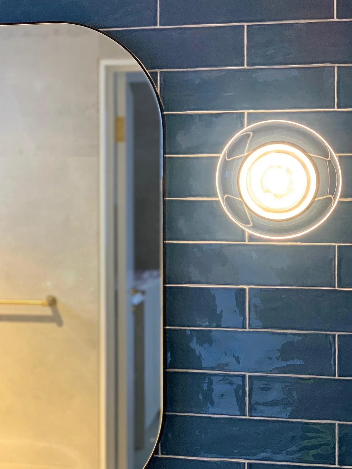 Bathroom mirror reflecting a door, mounted on a wall with dark blue tiles, illuminated by a round ceiling light.
