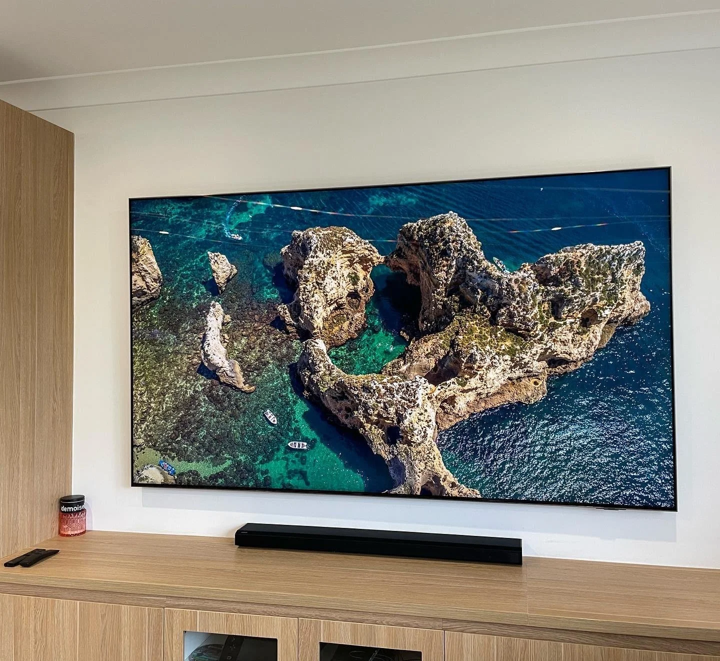 Large flat-screen TV mounted on a white wall displays an aerial view of a rocky coastline with turquoise water and small boats. A soundbar is placed below the TV, and to the left, there is a container and a remote control on a wooden cabinet.