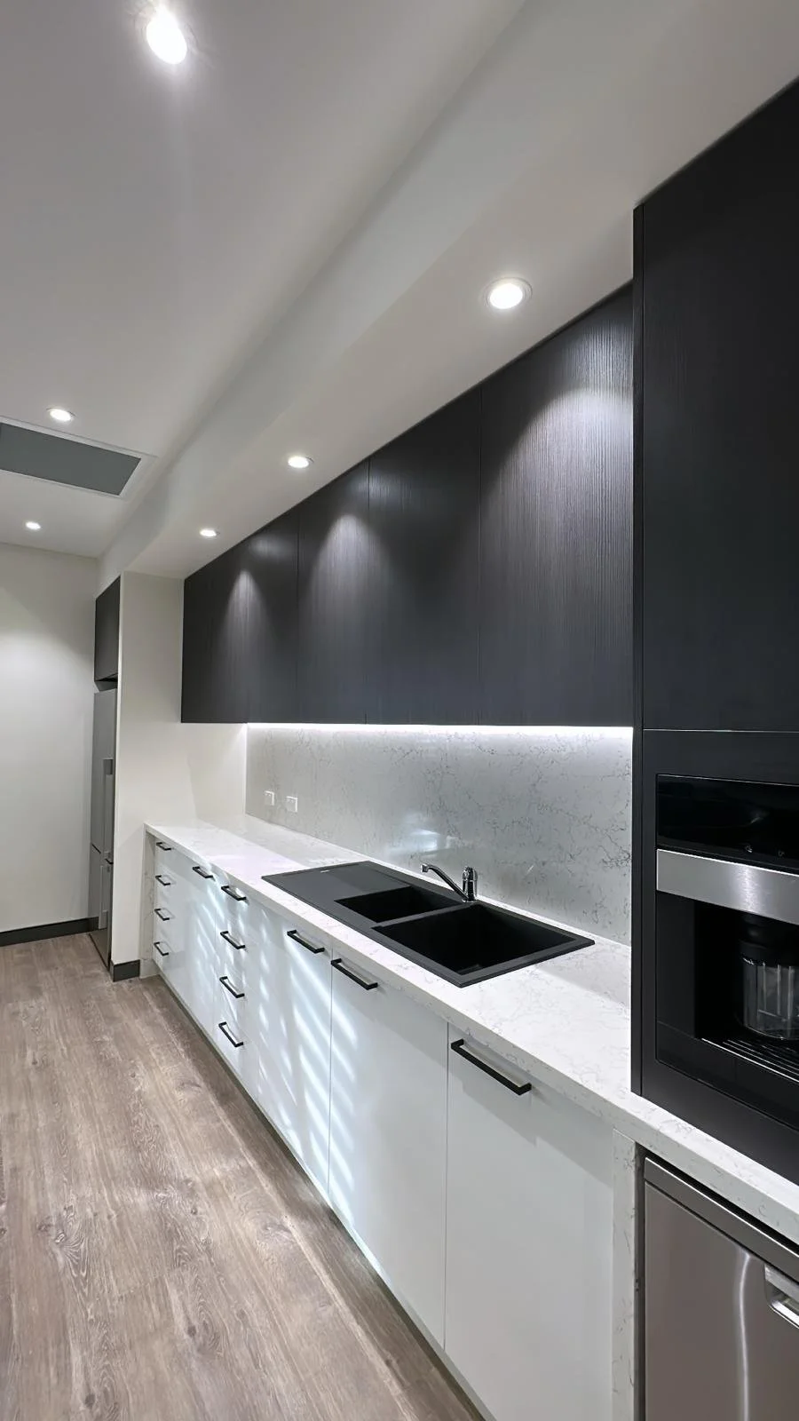 Modern kitchen with black upper cabinets, white lower cabinets, marble backsplash, black sink, and built-in appliances, illuminated by recessed ceiling lights.