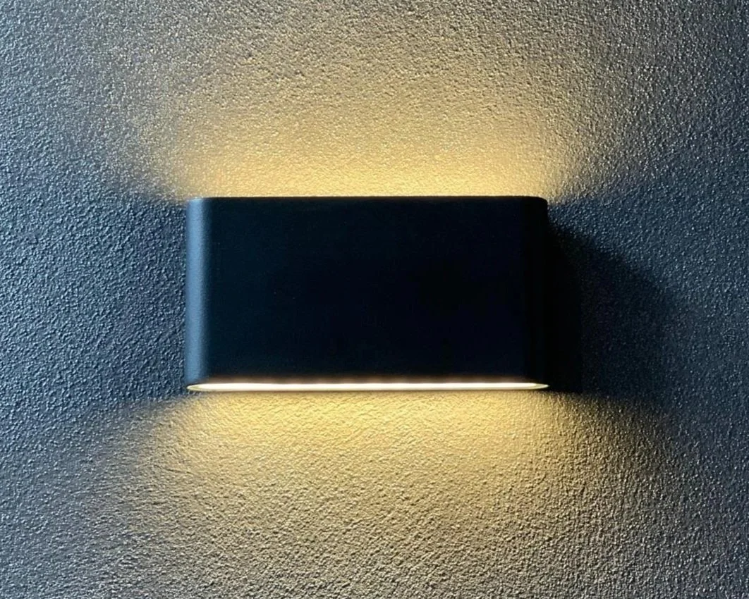 Modern wall sconce light fixture with a black rectangular cover emitting warm light onto a textured wall.