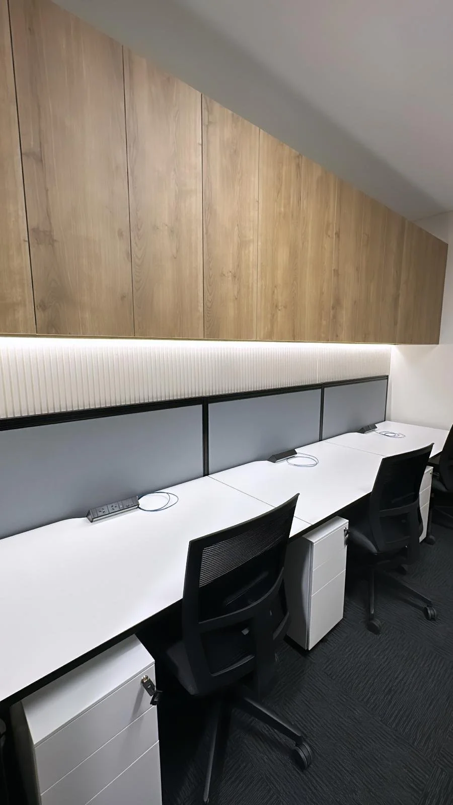 Office cubicles with three workstations, black chairs, white desks, power outlets, and a wooden wall with lighting above.