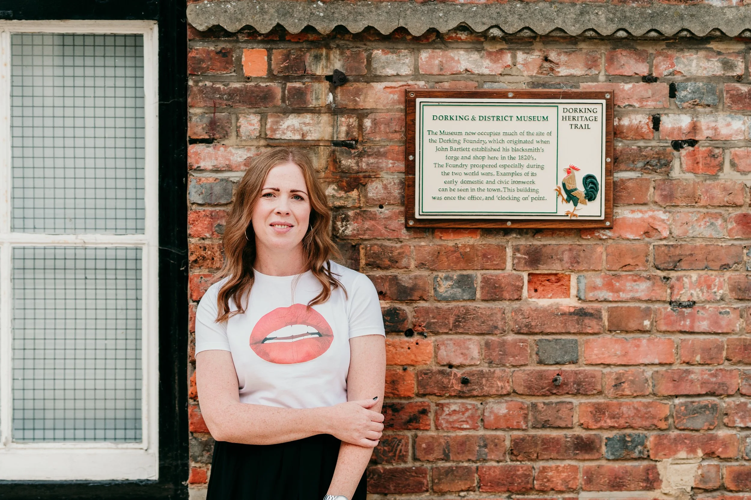 A woman with shoulder-length brown hair, wearing a white t-shirt with a red lips graphic, stands in front of a brick wall beside a window with a white frame and grid pattern, and a sign about the Dorking & District Museum.