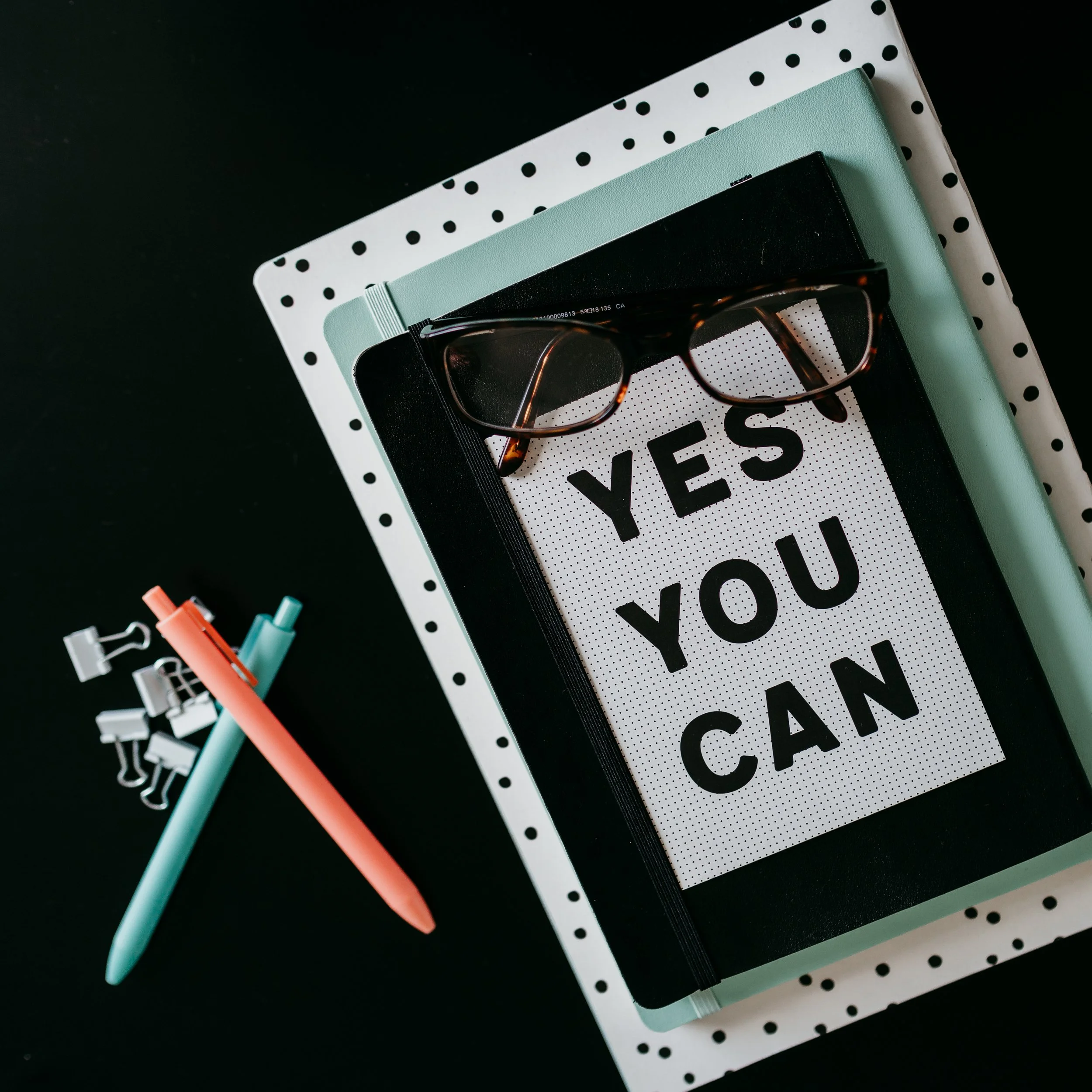 A black notebook with a pair of glasses resting on top, placed on a white polka-dotted tray, with two pens and small clip notes nearby, and a sign reading 'YES YOU CAN' underneath.