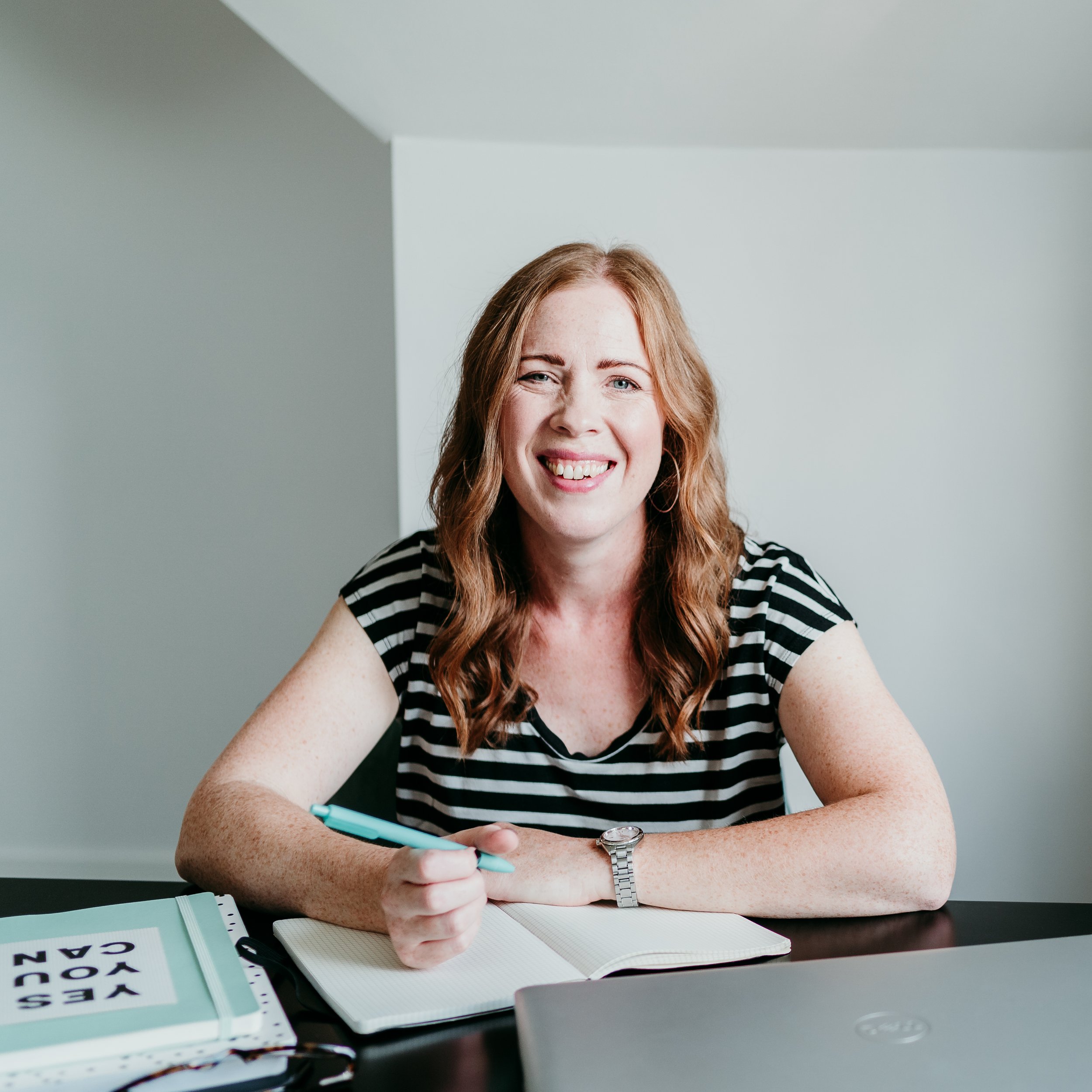 A woman with red hair and freckles, wearing a black and white striped shirt, sitting at a desk with a notebook, a teal pen, and a binder that reads 'YES YOU CAN,' smiling at the camera in a well-lit room.