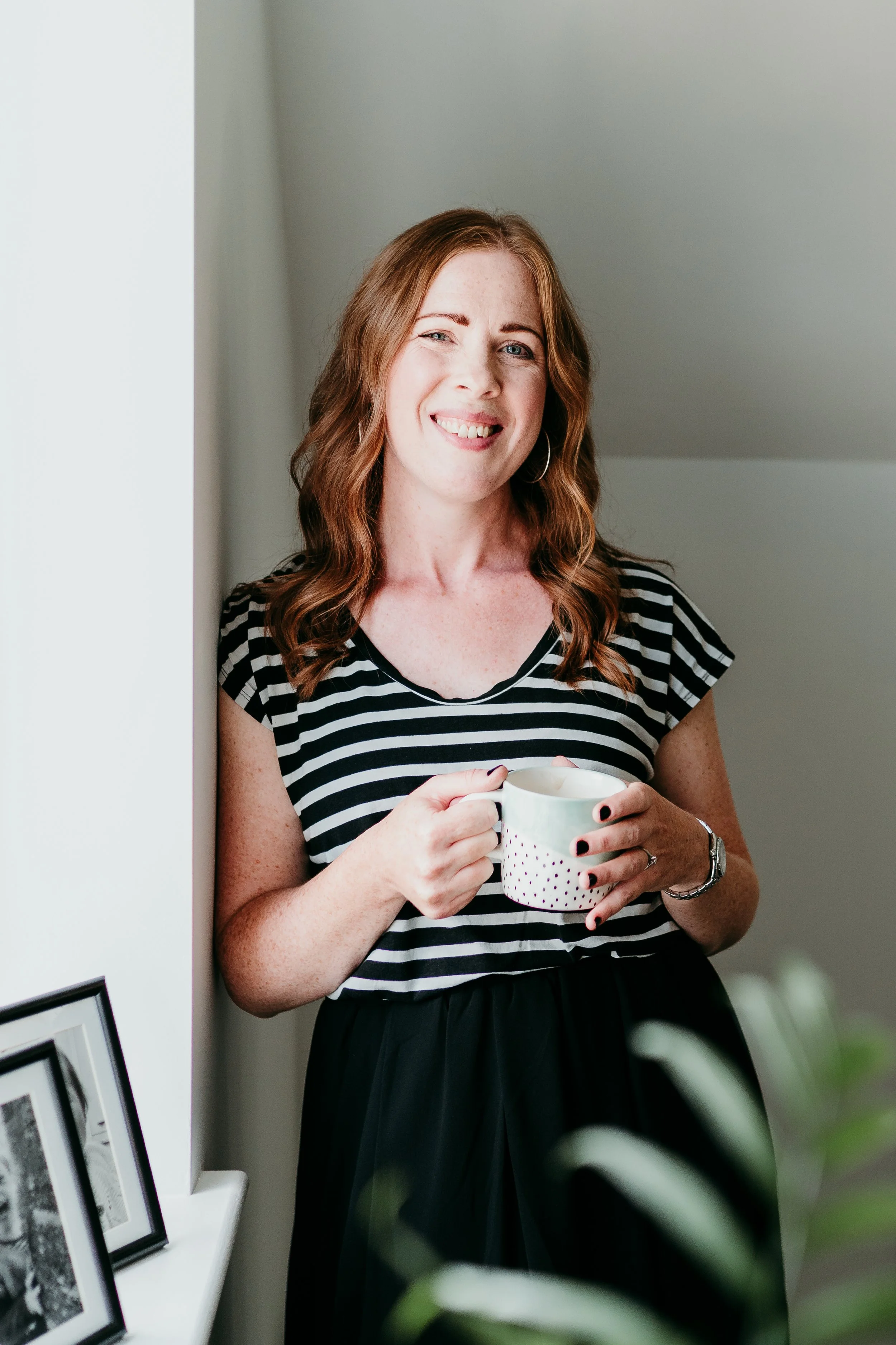 A woman with red hair, wearing a striped black and white shirt, holding a mug, standing indoors, smiling at the camera.