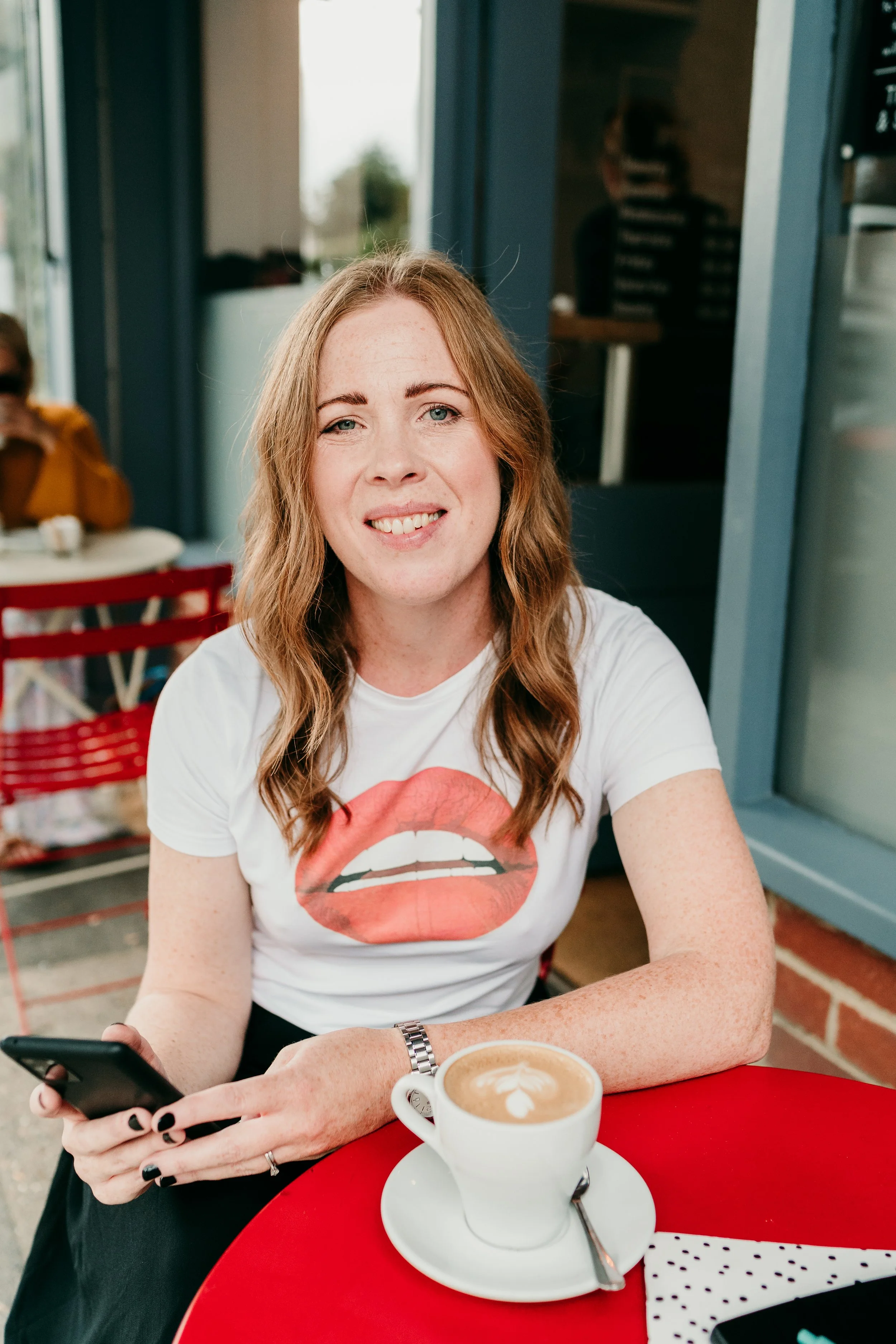 A smiling woman with red hair sitting at a red table in a cafe, holding a phone, with a cup of latte decorated with leaf art in front of her.