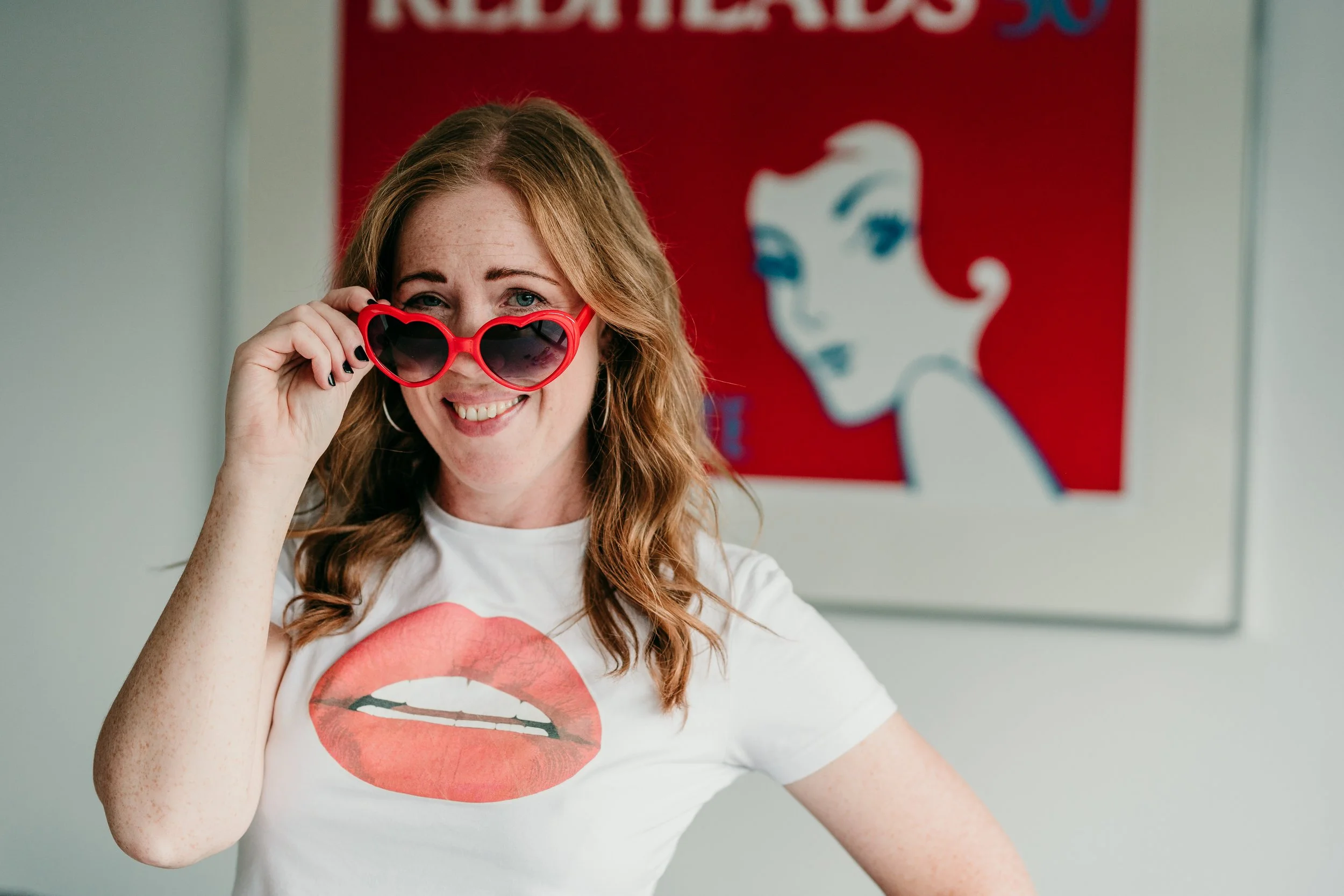 A woman with red hair wearing sunglasses and a graphic t-shirt with a lipstick kiss mark, smiling at the camera with a retro-style portrait of a woman in the background.
