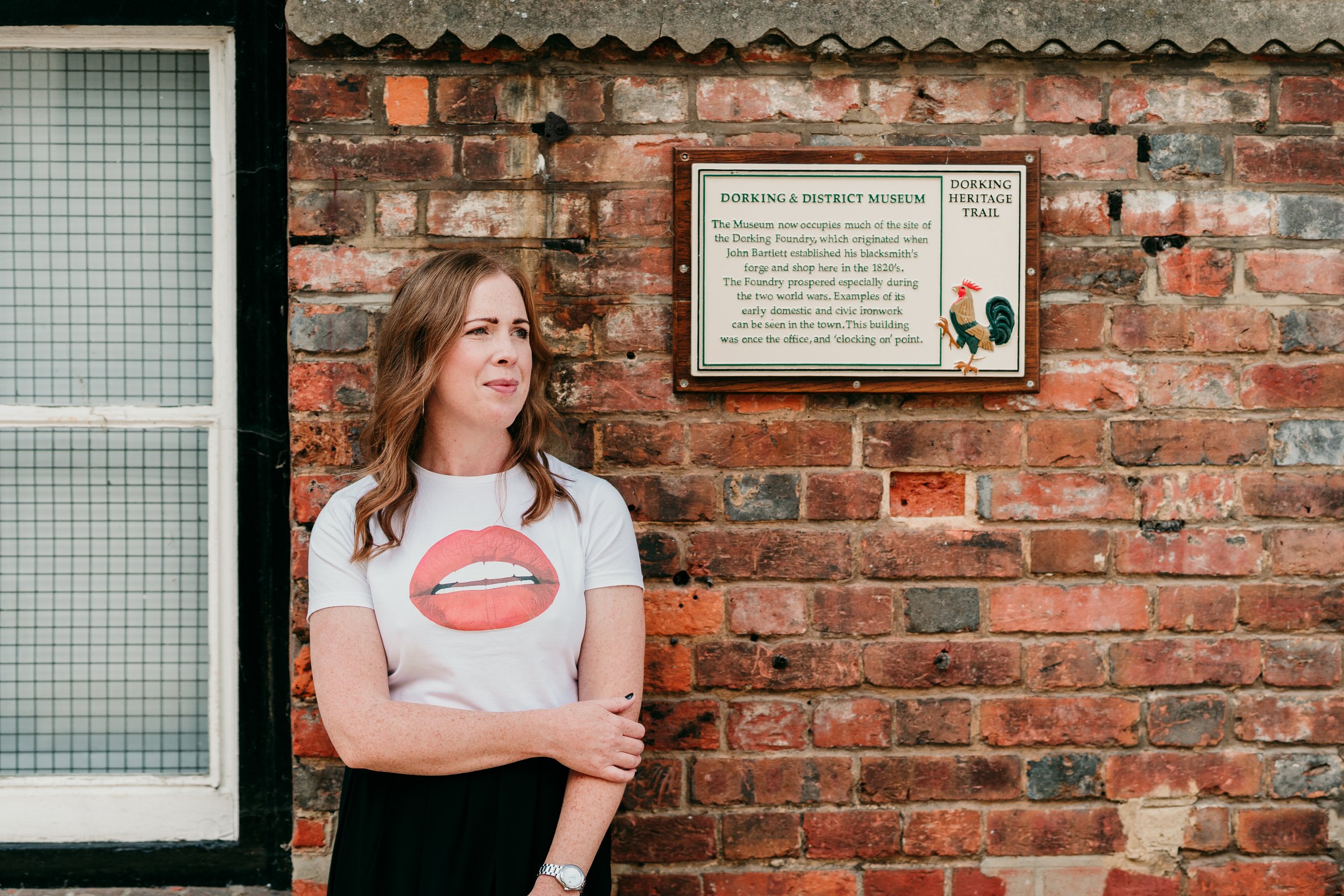 A woman with brown hair wearing a white t-shirt with a red lips graphic stands outside next to a brick wall with a sign about the Dorking & District Museum and a window.